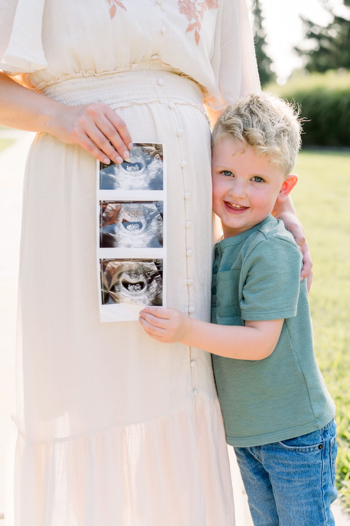 A young boy hugs his pregnant mother outdoors, holding ultrasound photos against her baby bump in a sunny park.