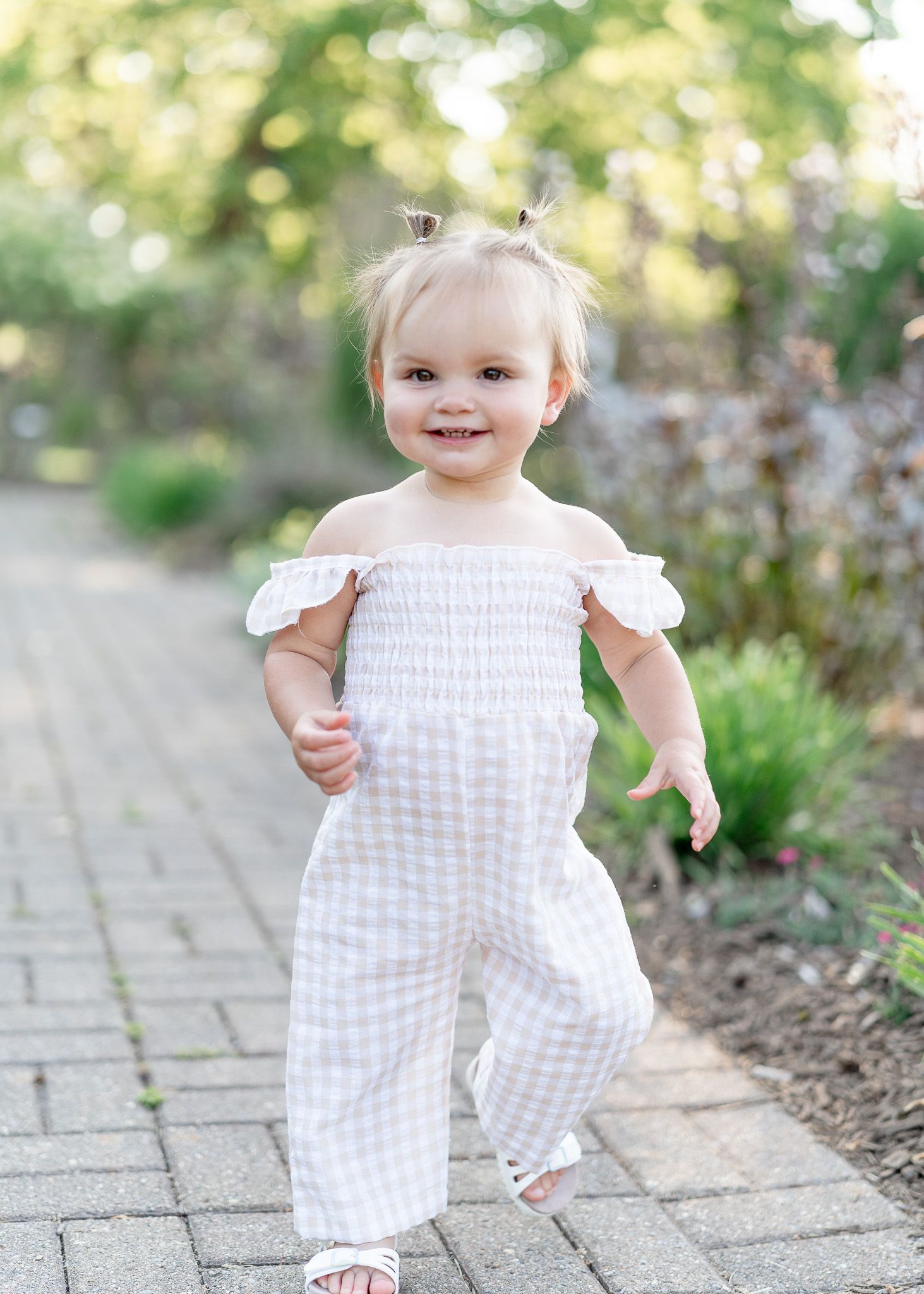 A small child wearing a white gingham off-shoulder romper stands on a stone pathway in a garden setting.