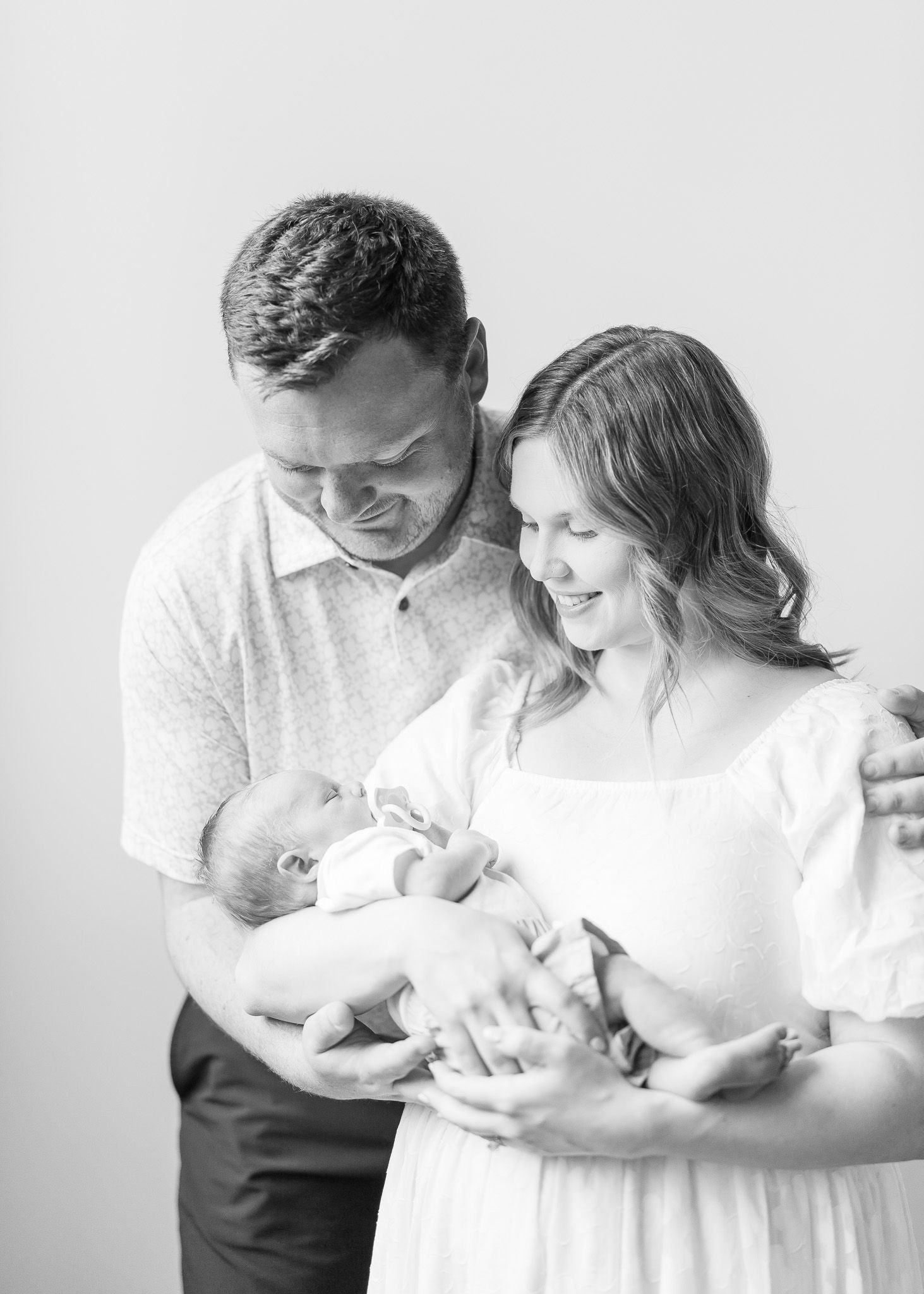 A tender black and white family portrait shows parents gazing lovingly at their newborn baby in a bright studio setting.