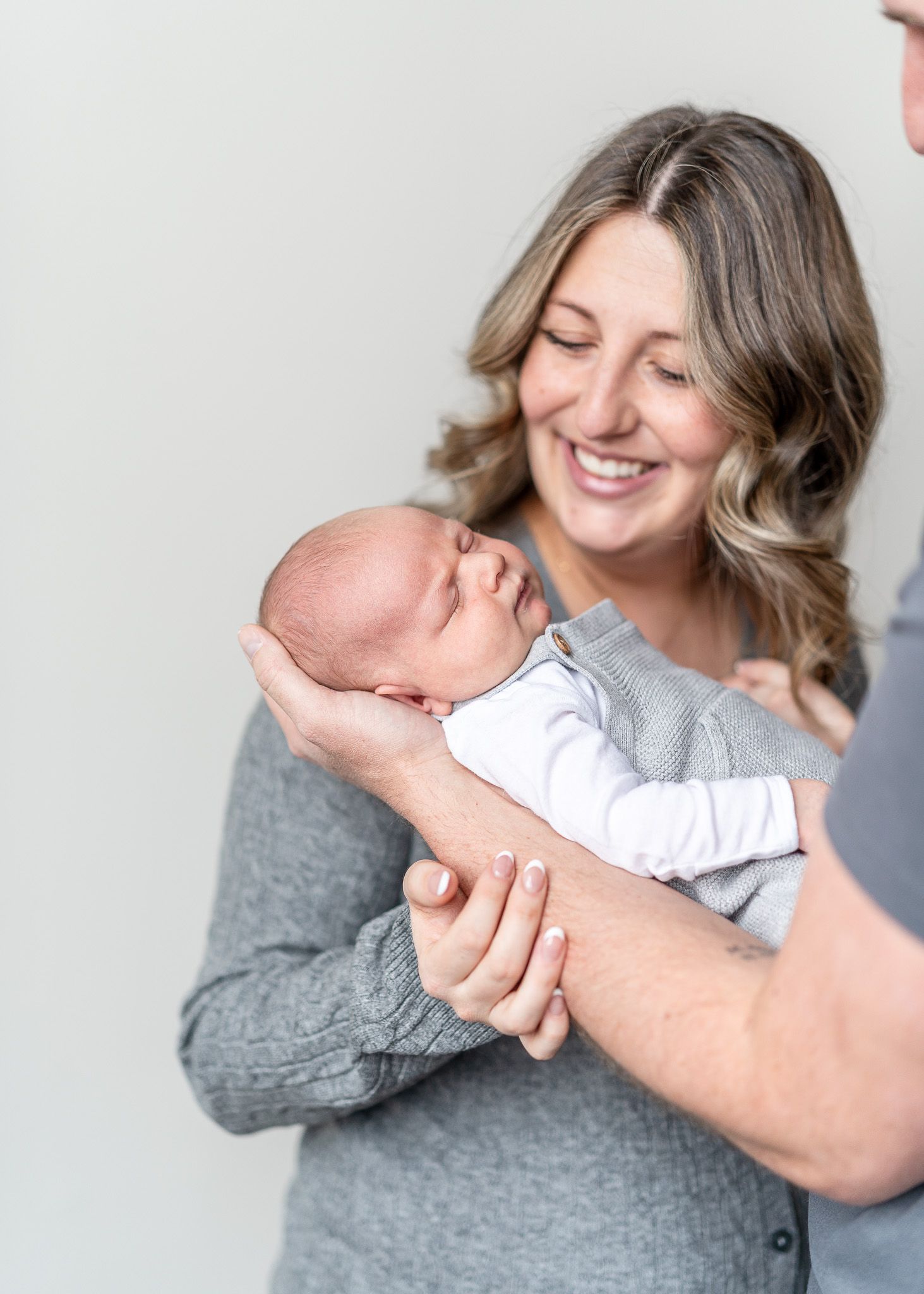 A tender moment captured as a woman in a gray sweater holds and smiles at a sleeping newborn baby in white.