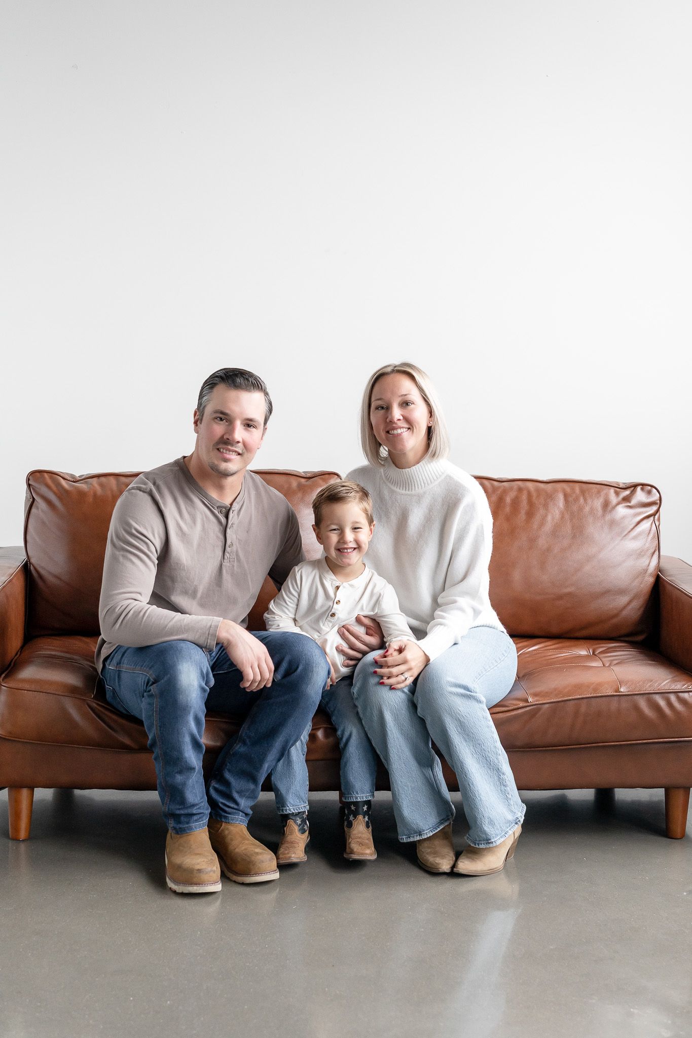 A family sits together on a brown leather sofa in a minimalist room with concrete floors.