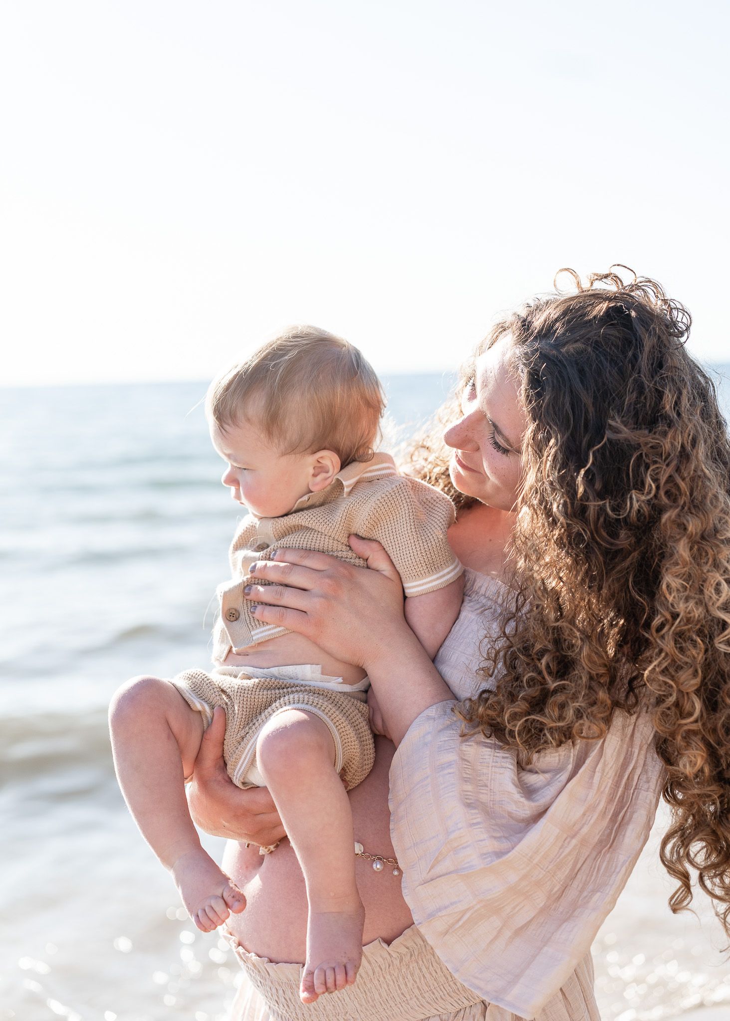 A parent and baby share a tender moment together on a sunny beach with waves in the background.
