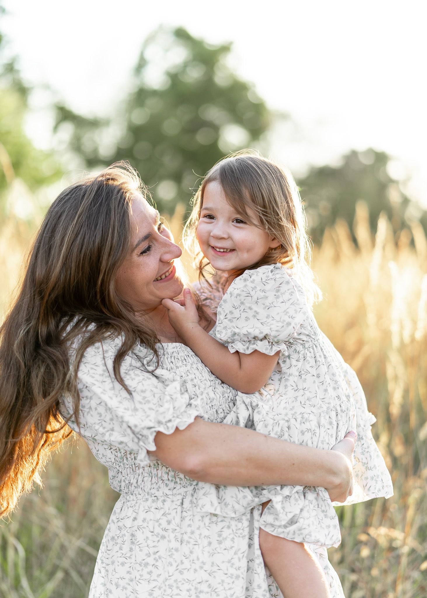 A tender moment shared between parent and child wearing matching white lace dresses in a sunlit field of tall grass.