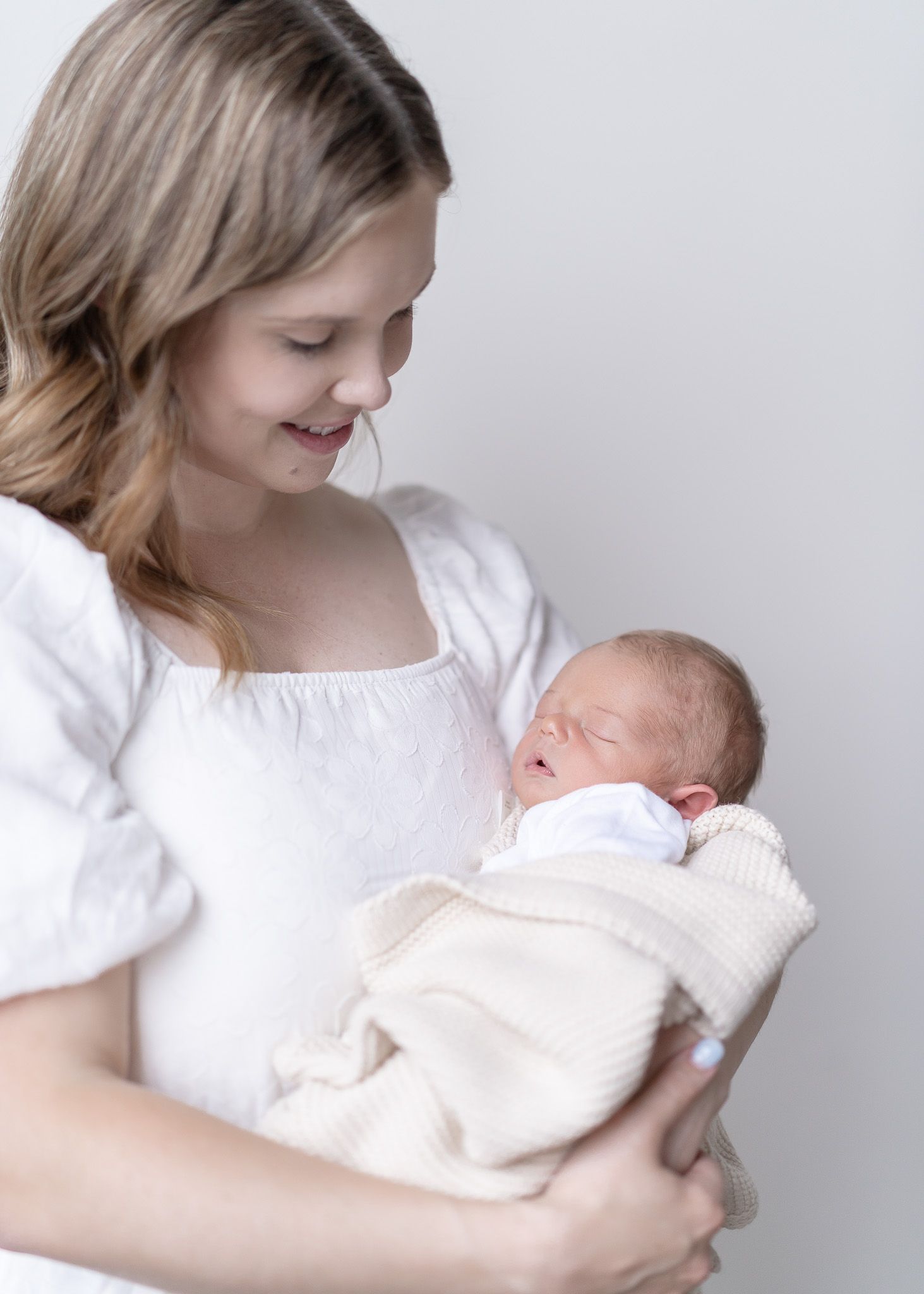 A mother in a white shirt tenderly holds and gazes at a newborn baby wrapped in a white blanket.