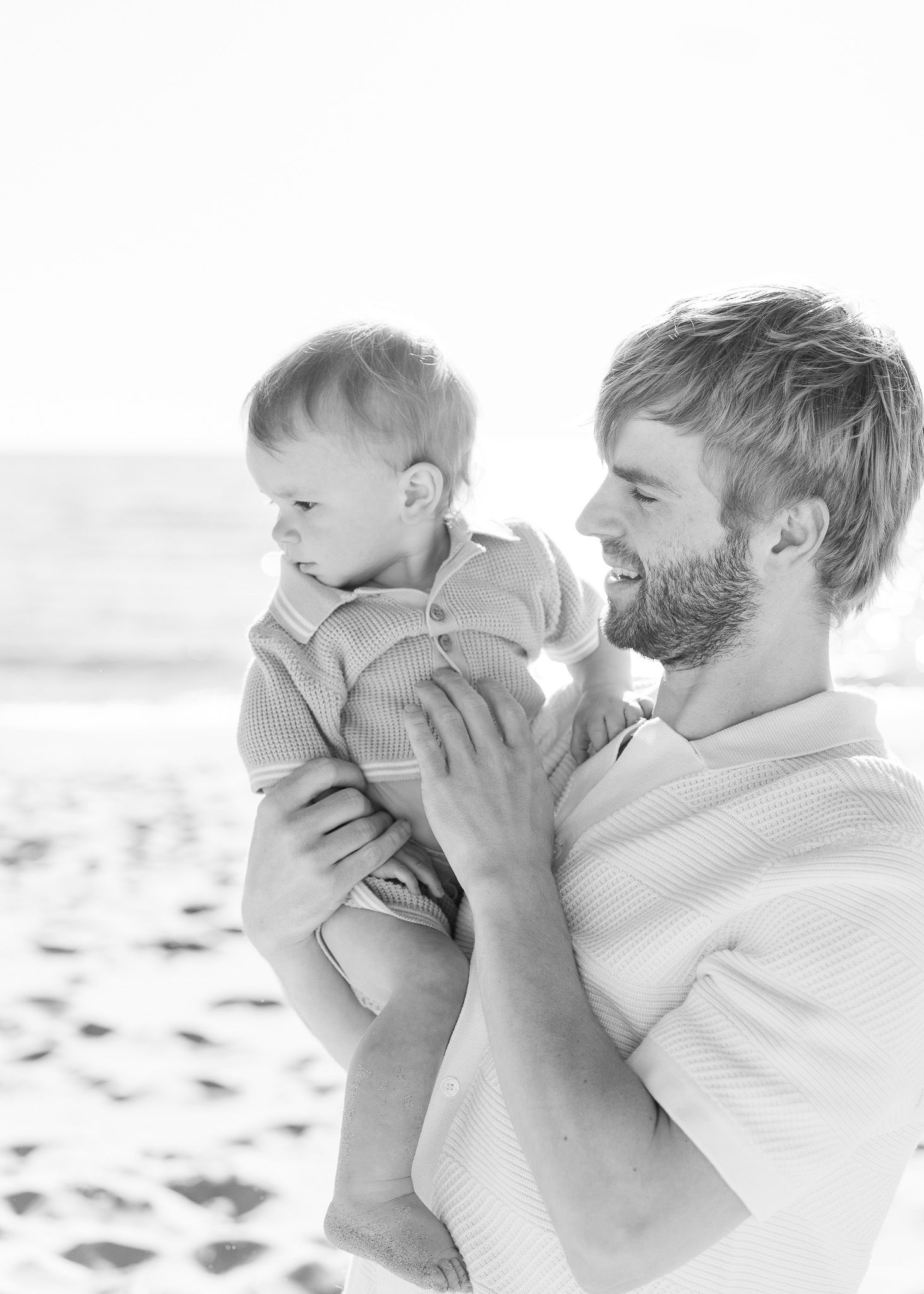 A tender black and white moment captured on the beach between parent and baby in casual attire.