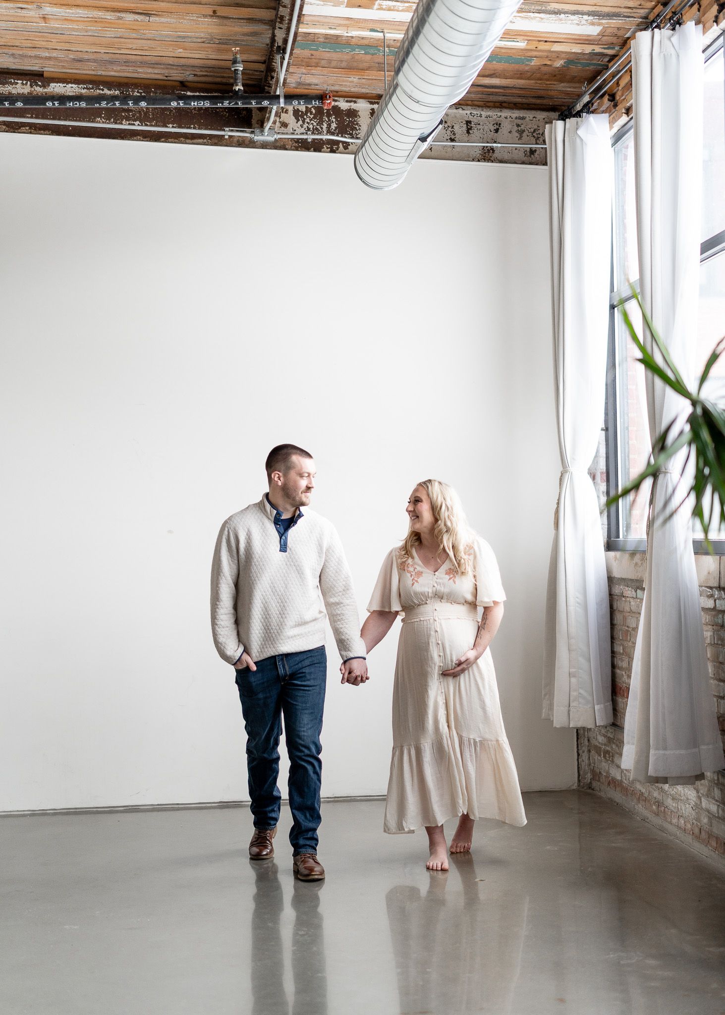 A couple dances together in a white industrial studio space with exposed wooden beams and white curtains.