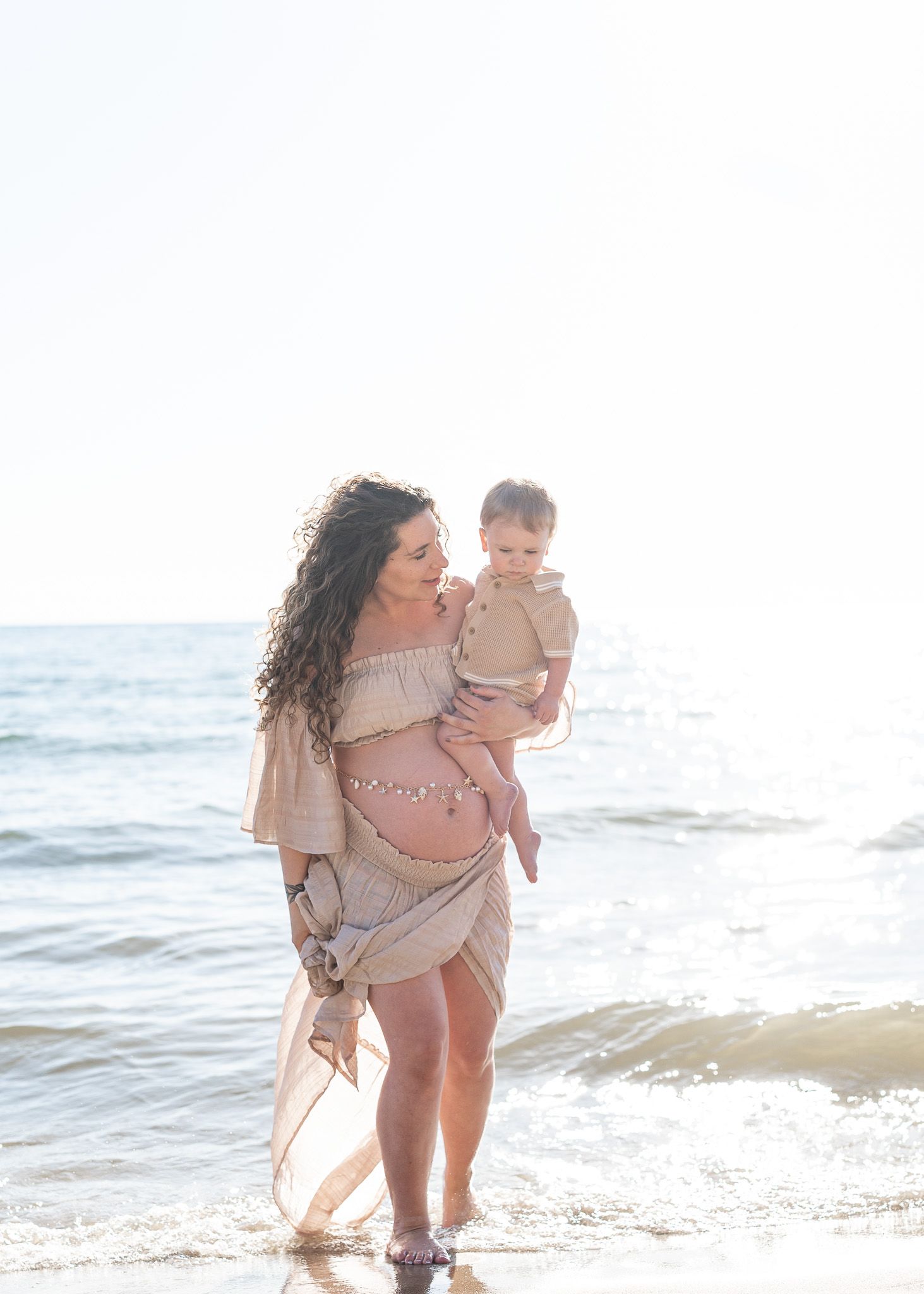 A pregnant woman in a flowy beige dress holds a toddler while standing on a sunny beach by the ocean waves.