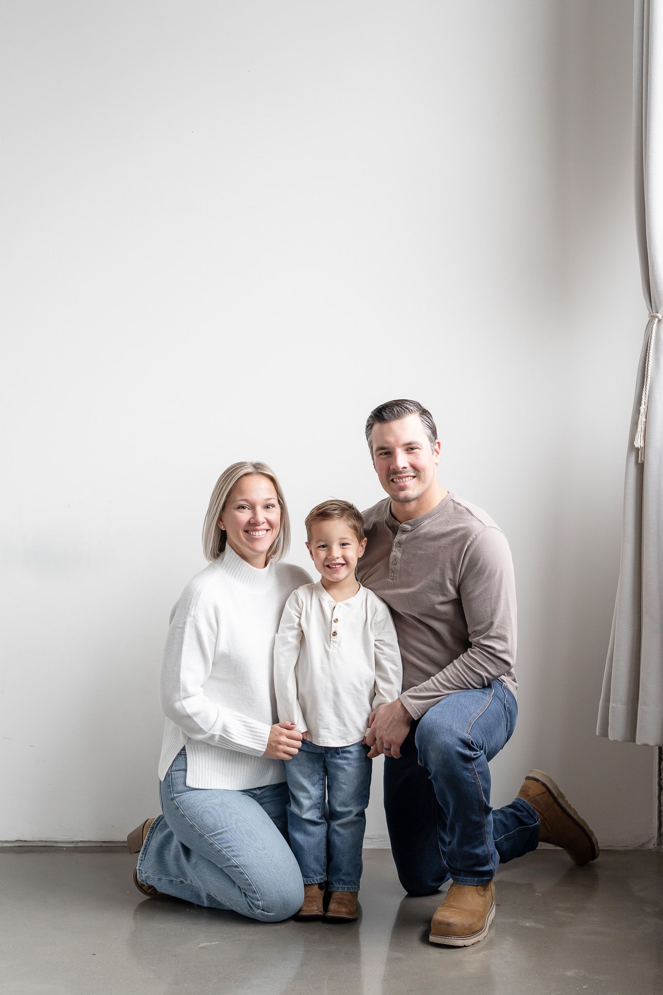 A family sits together in a minimalist white room wearing casual clothing and jeans while smiling and laughing.
