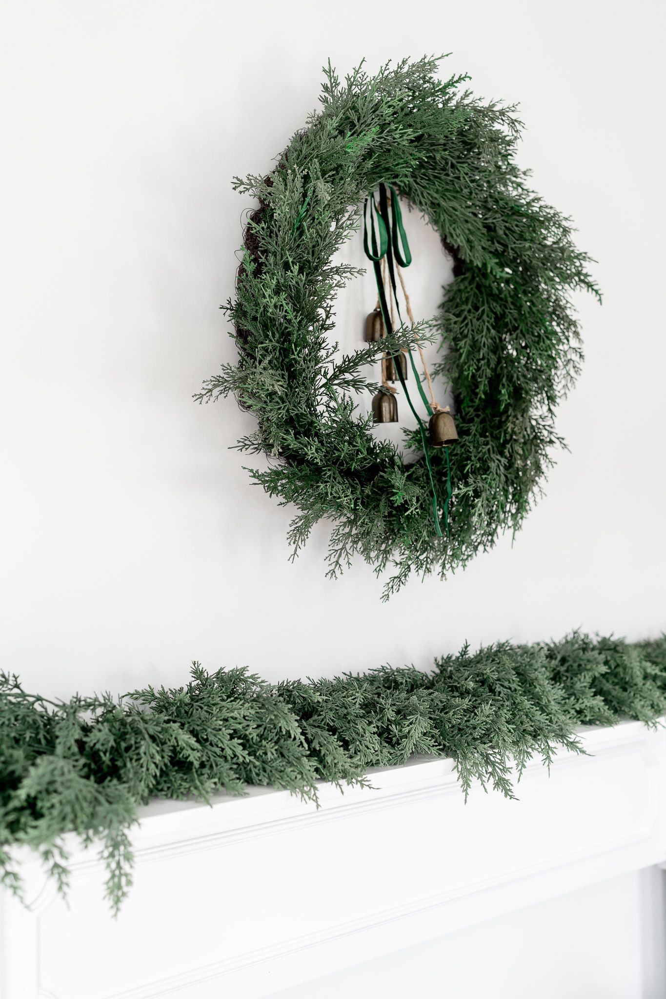 A fresh evergreen wreath and garland adorned with bells hangs on a white wall.