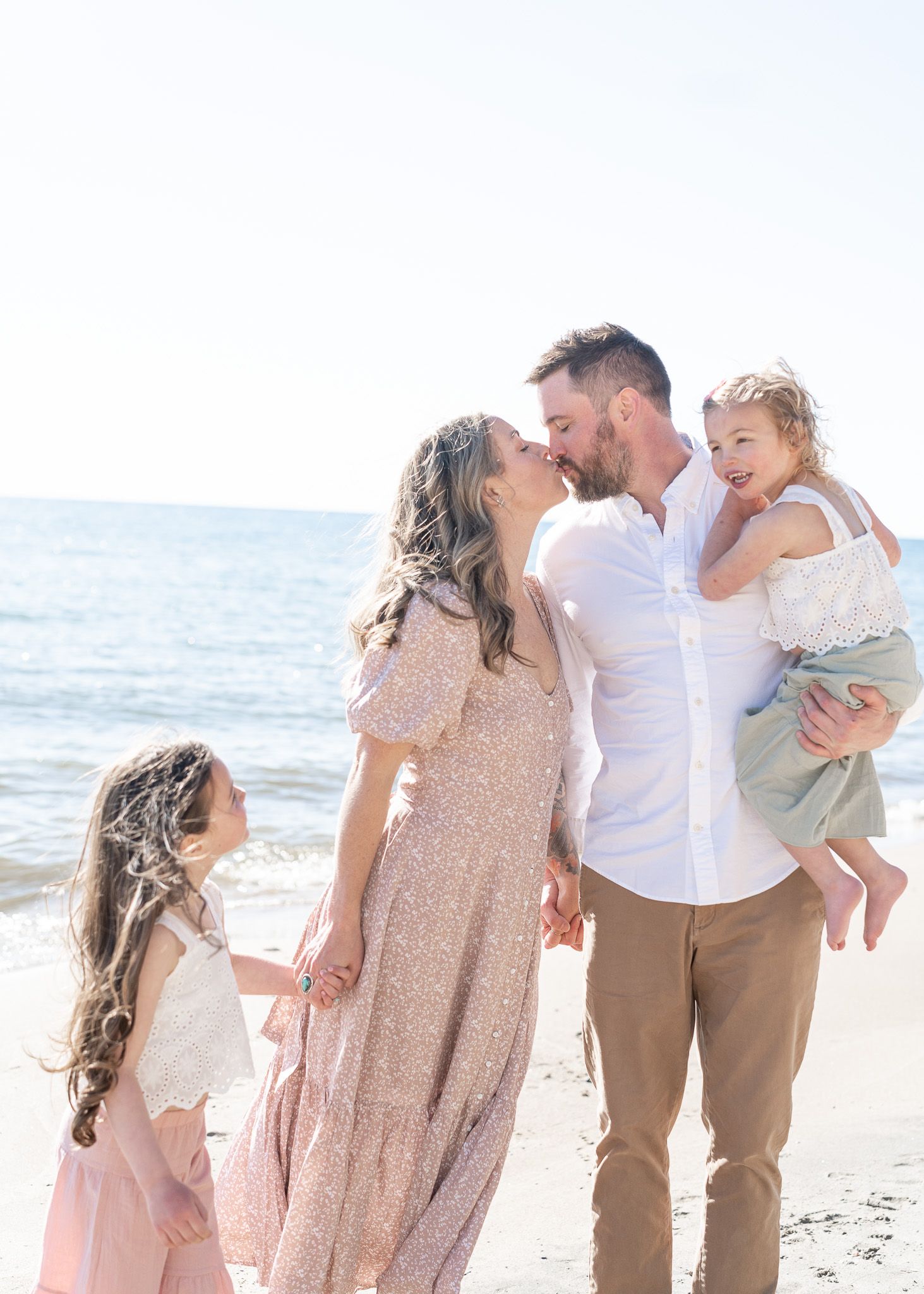 A family in coordinating beige and white outfits shares tender moments together on a sunny beach.