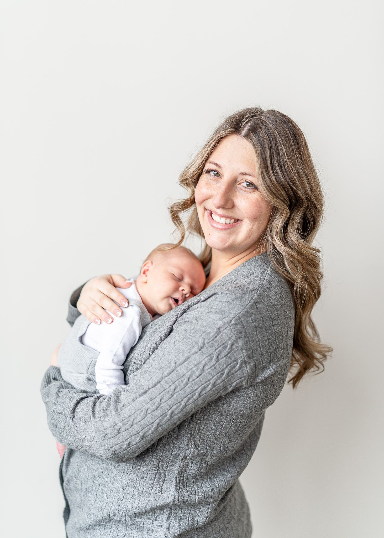 Mother in gray sweater lovingly embraces and comforts sleeping baby in white outfit against light background.