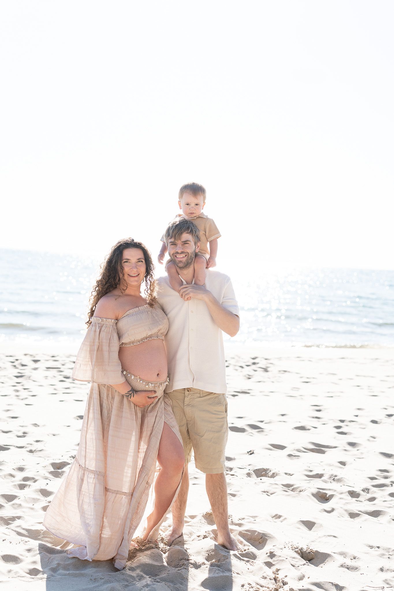 Family wearing light colored clothing embraces while standing on a sunny beach with ocean waves in background.