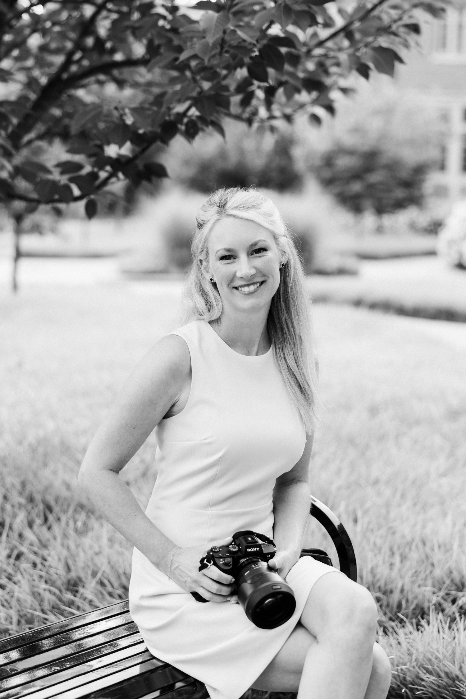 Black and white portrait of a smiling blonde woman sitting on a bench in a park, holding a DSLR camera.