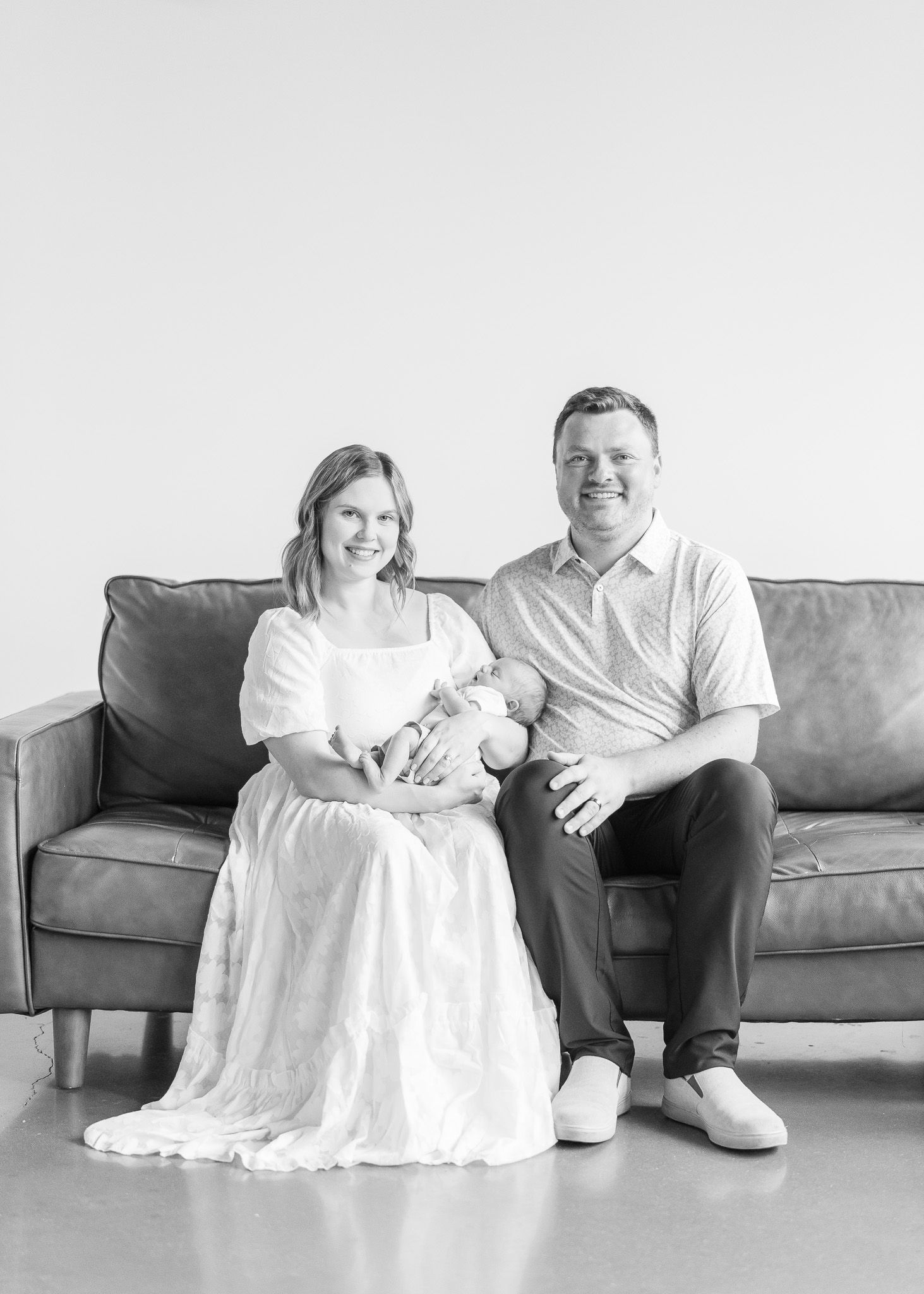 A couple in white attire sits on a gray couch, captured in three similar poses against a minimalist backdrop.