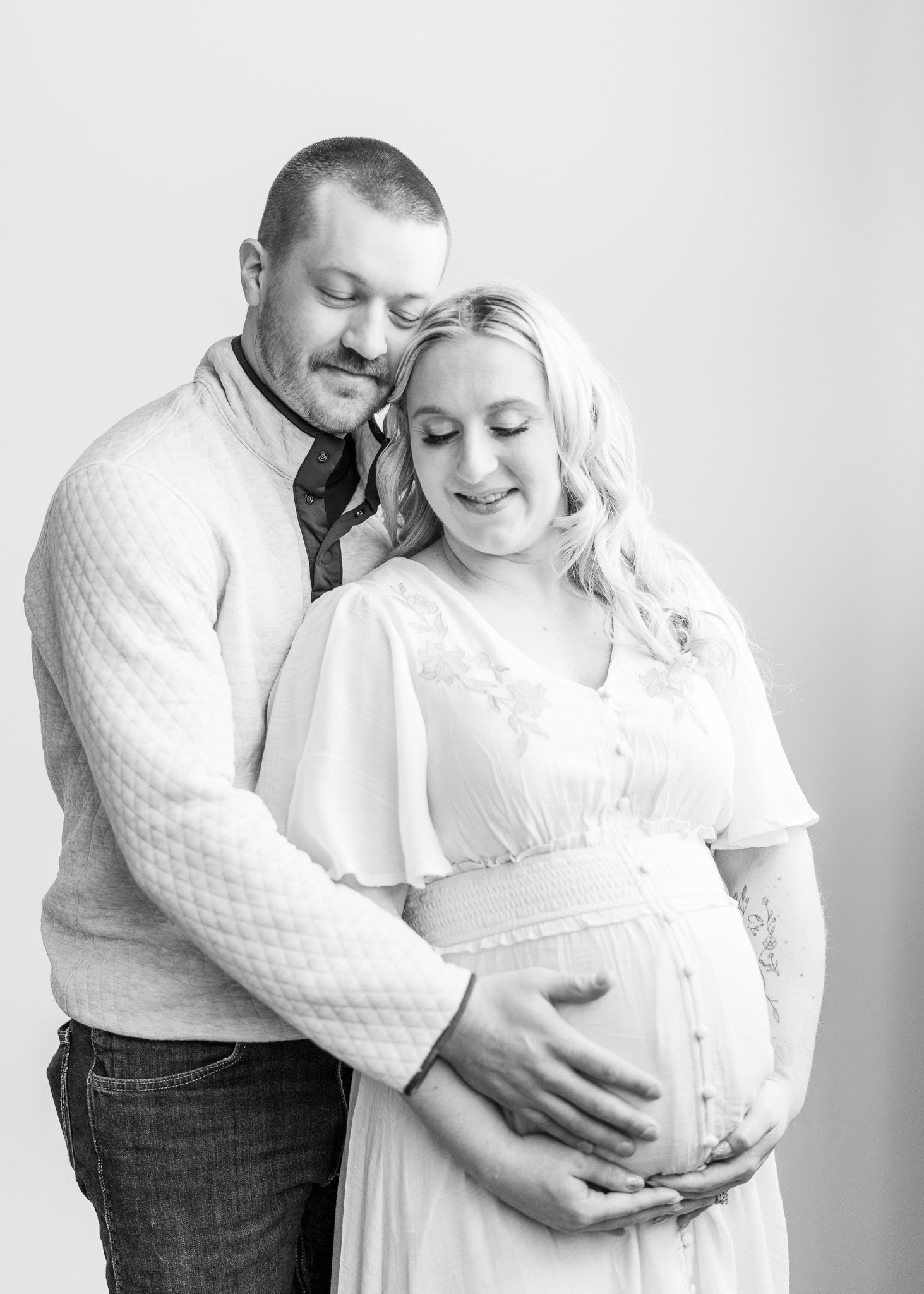 Black and white maternity photos showing tender moments between expectant parents in white clothing against neutral backdrop.