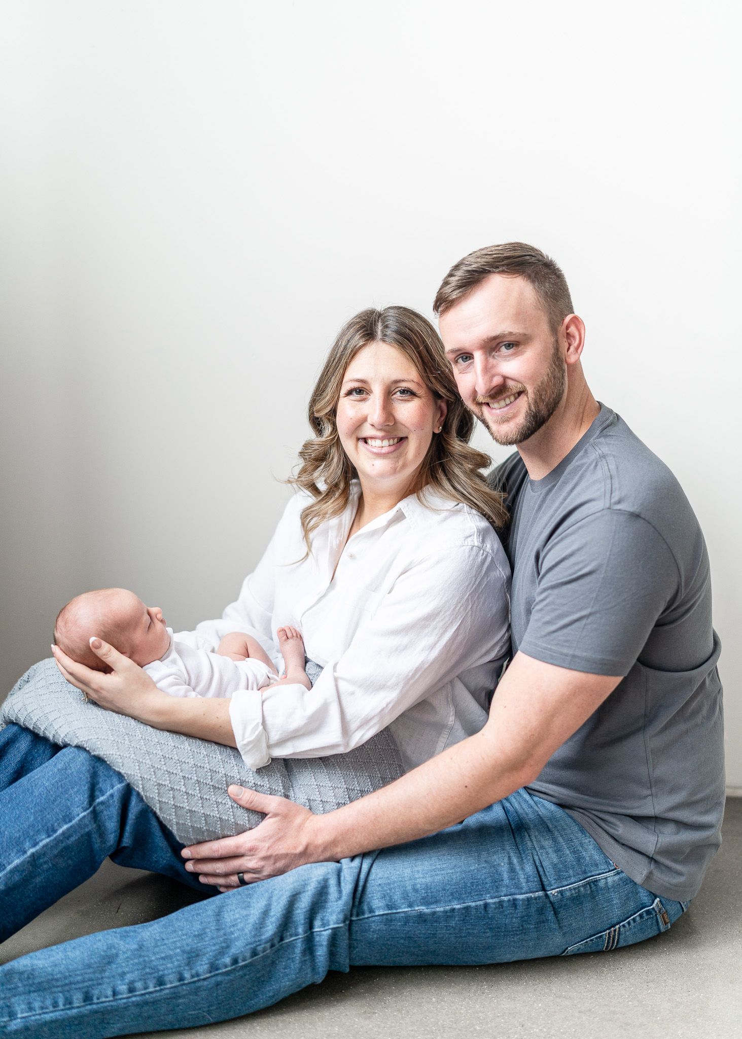 A couple in casual attire lounges together on a white floor in a series of intimate maternity photoshoot poses.