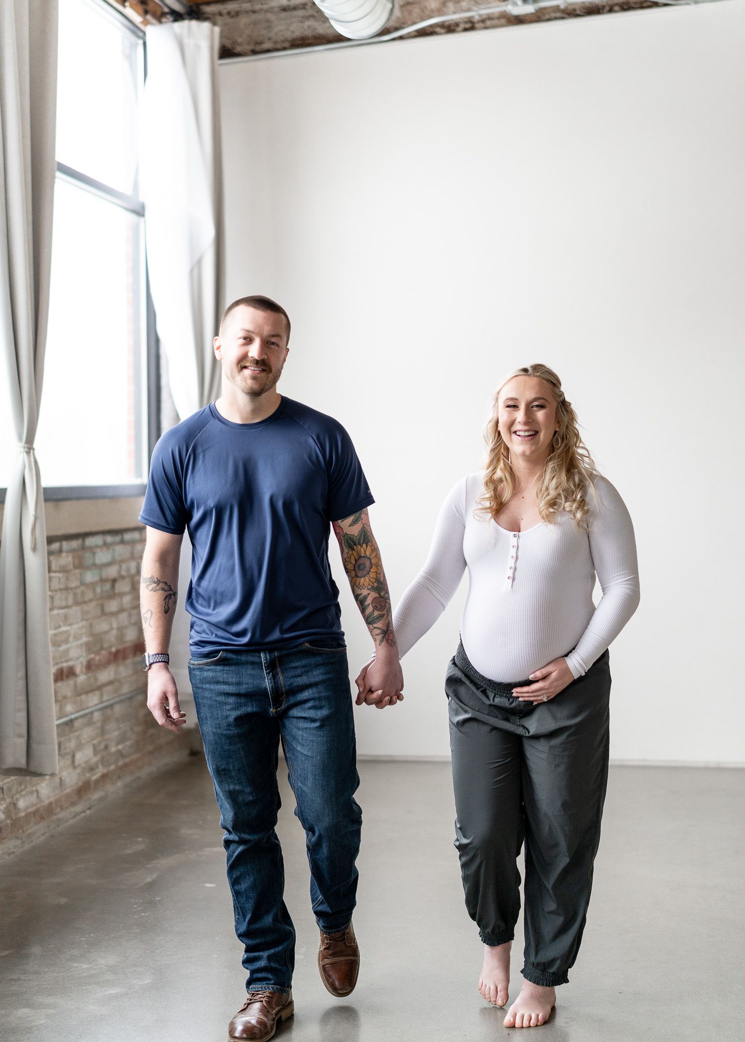 A couple holds hands while walking together in a bright industrial-style loft space with exposed brick walls.