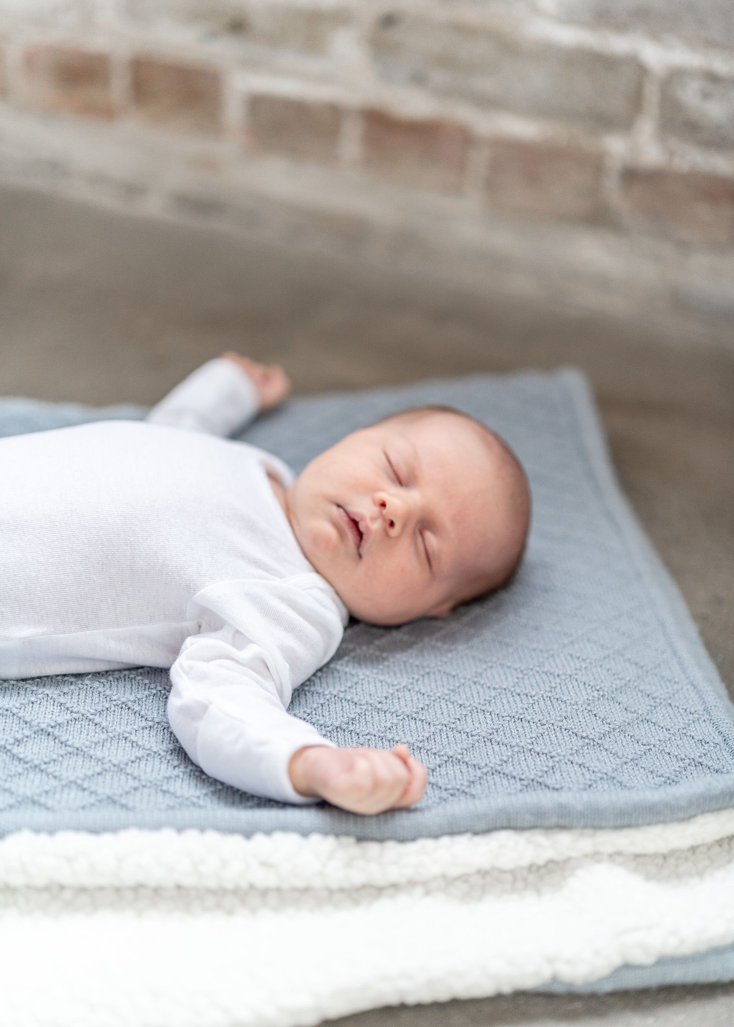 Peaceful sleeping newborn baby wearing white outfit rests on soft gray knitted blanket against brick wall background.