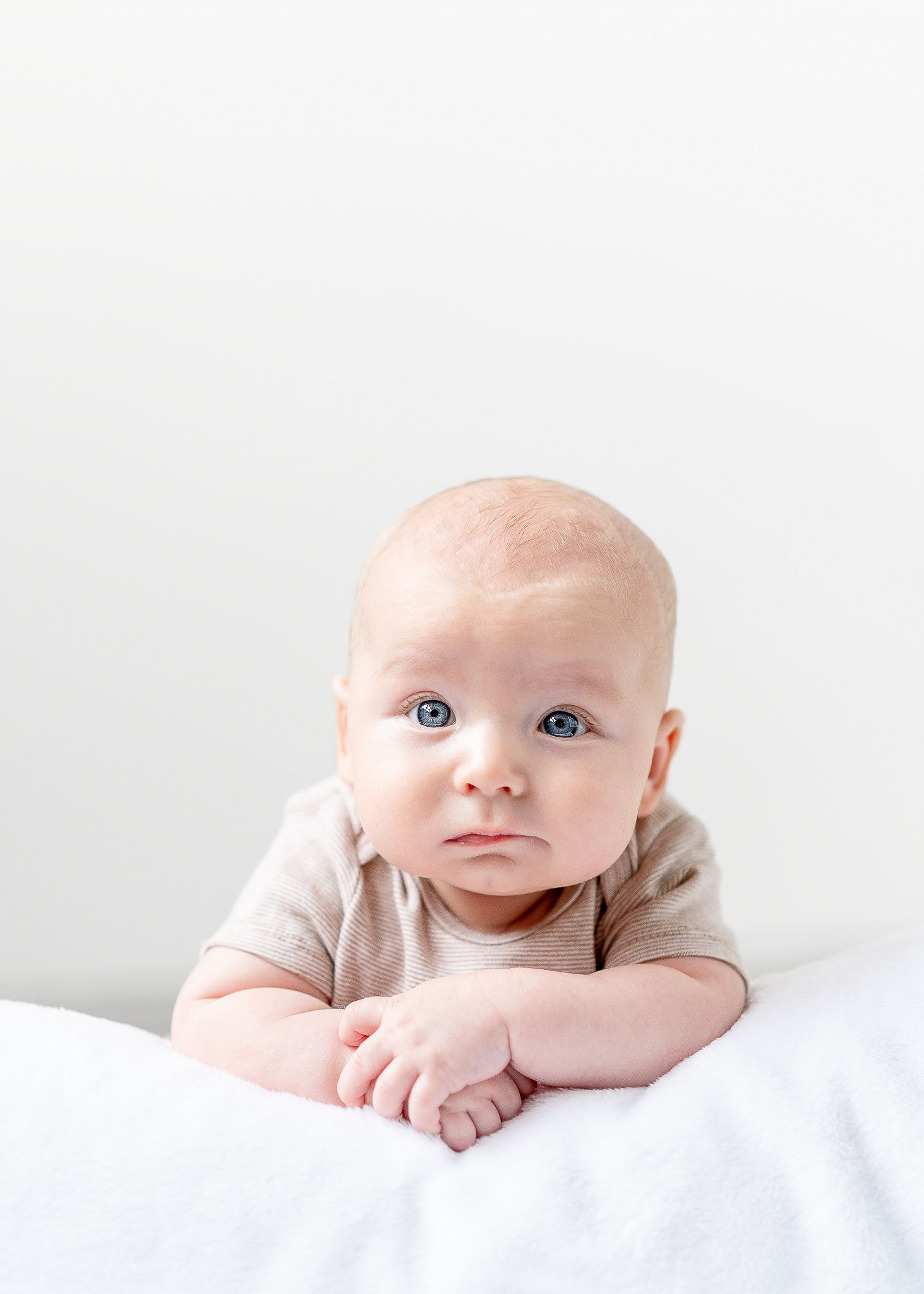 A baby lying on white bedding looks up with bright blue eyes and a gentle expression.