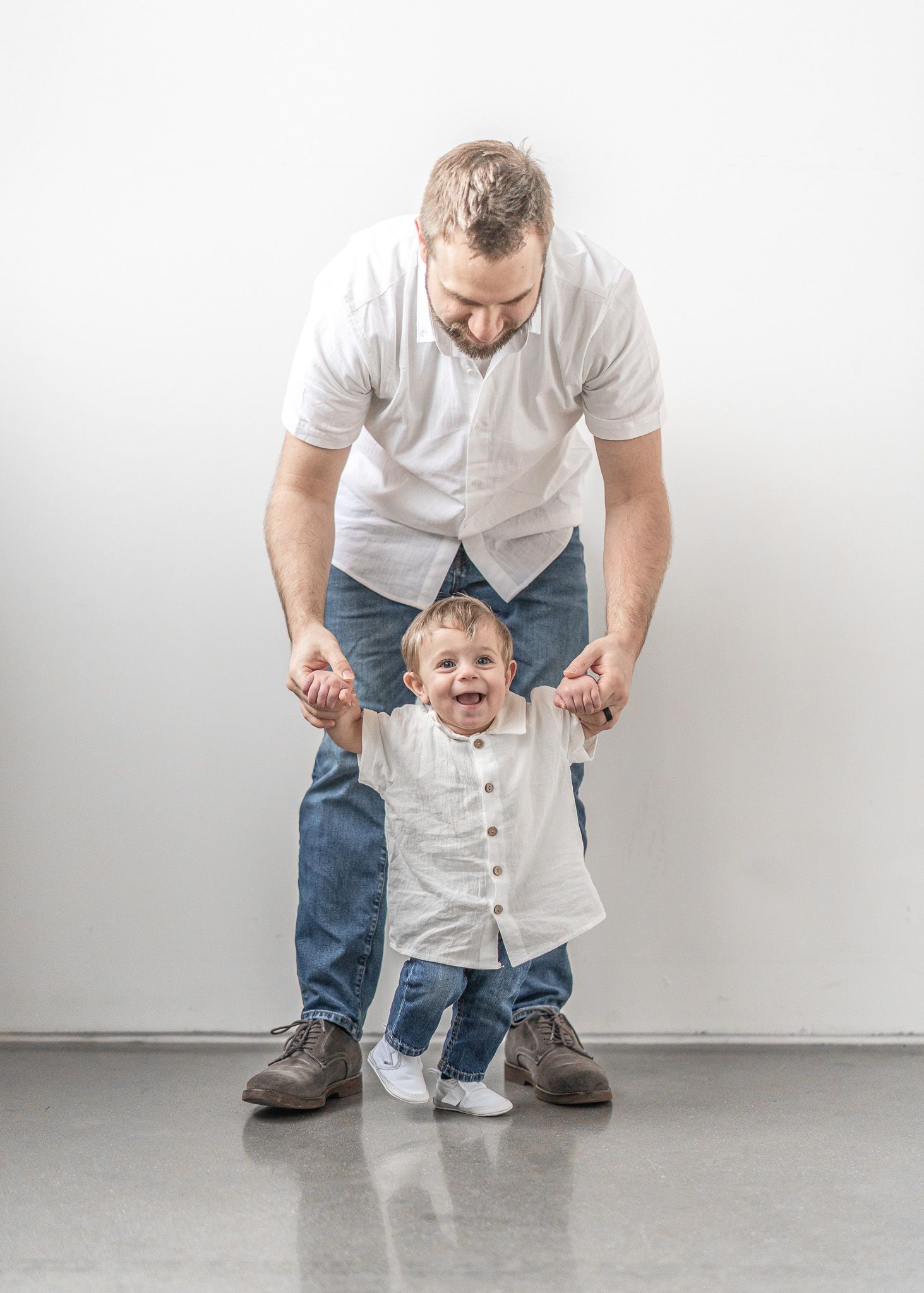 Parent in white shirt and jeans helps small child learn to walk on gray floor in minimalist setting.