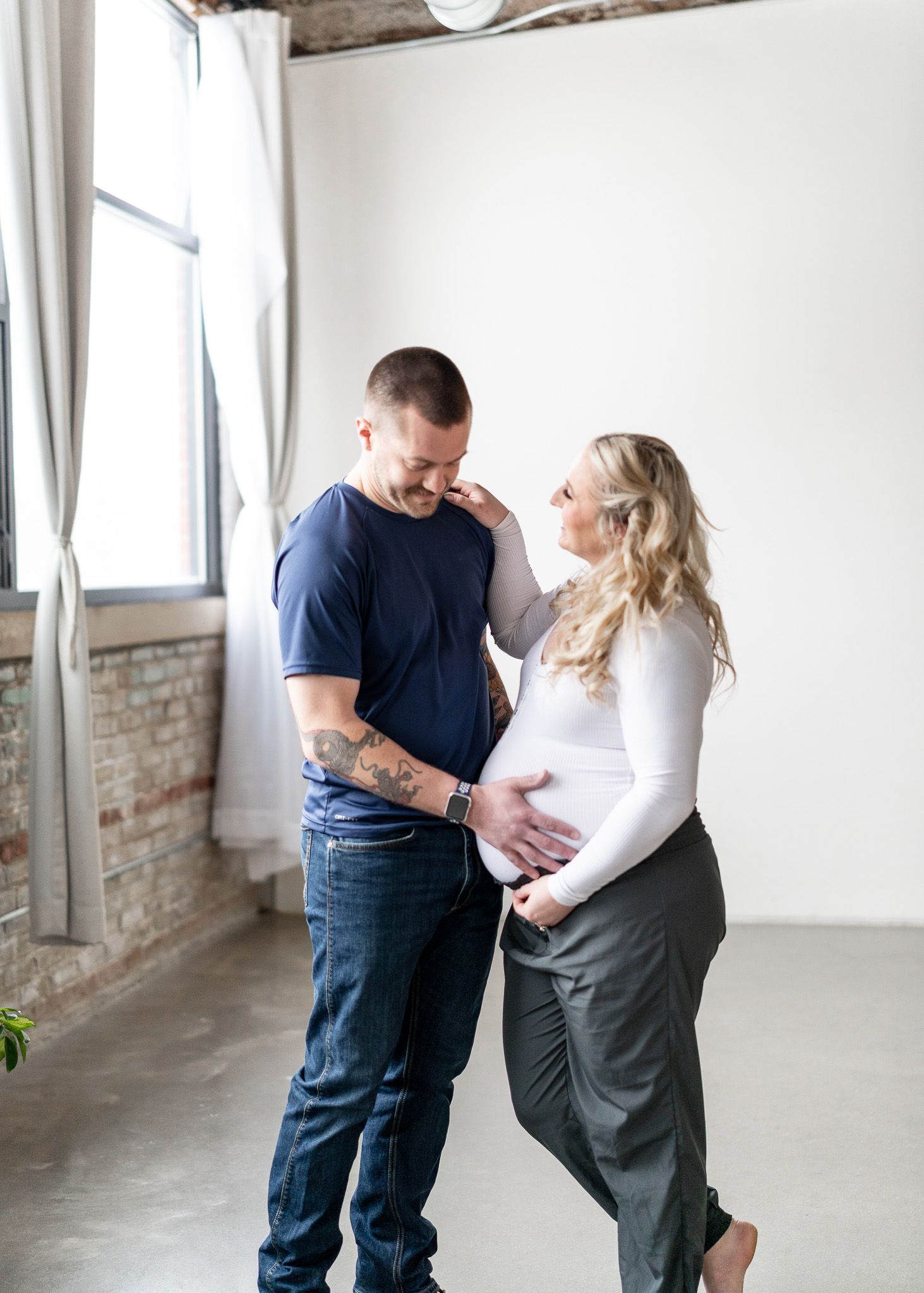 A couple shares a tender moment in a bright industrial loft space with exposed brick walls and white floors.