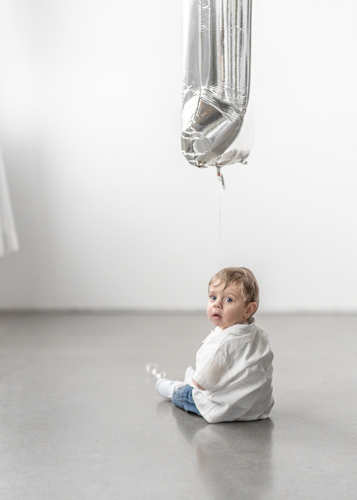 Small child in white sweater sits on grey floor looking up at silver mylar balloons in minimalist setting.