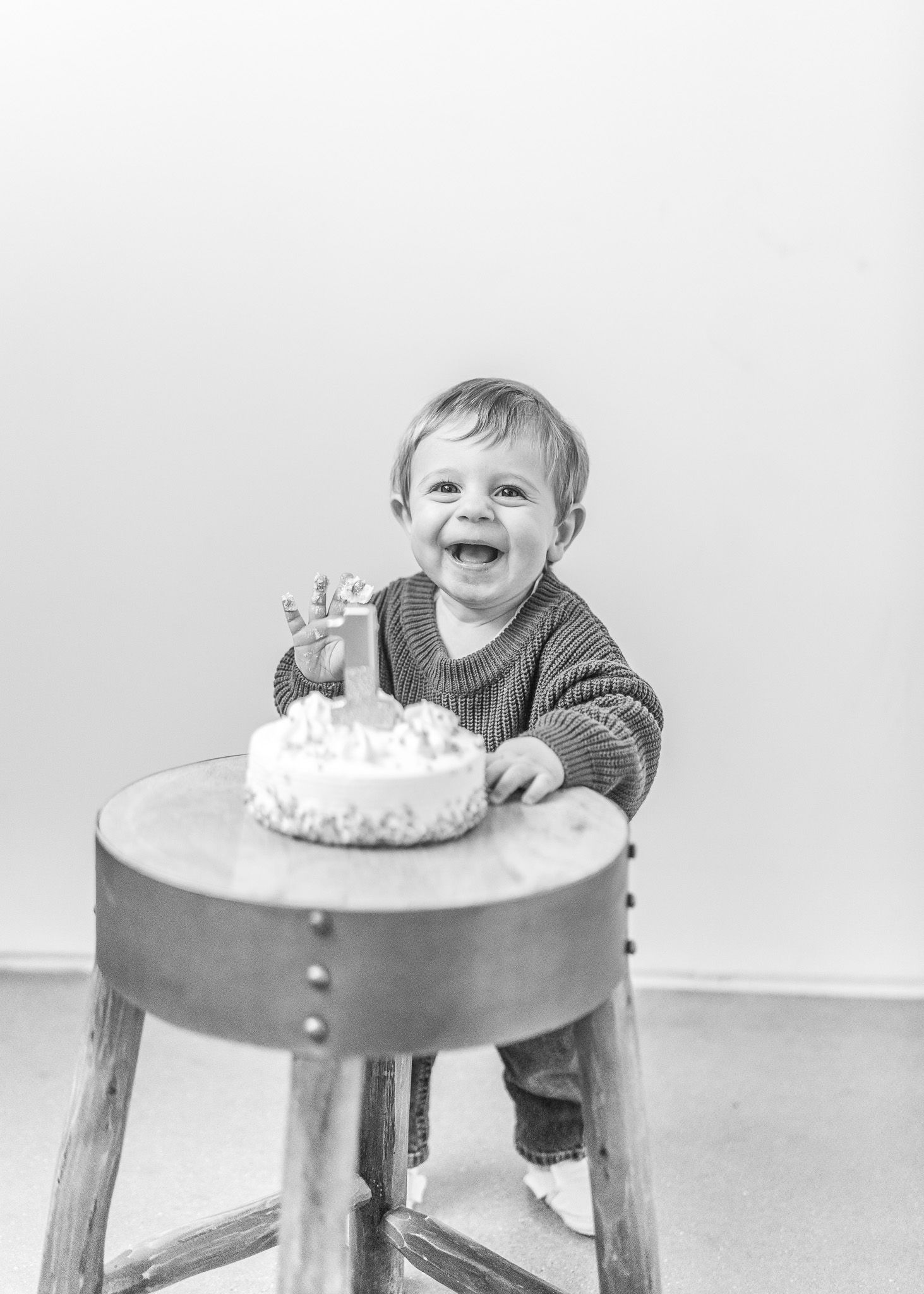 Four sequential photos show a baby joyfully smashing and playing with a white frosted birthday cake on a round table.