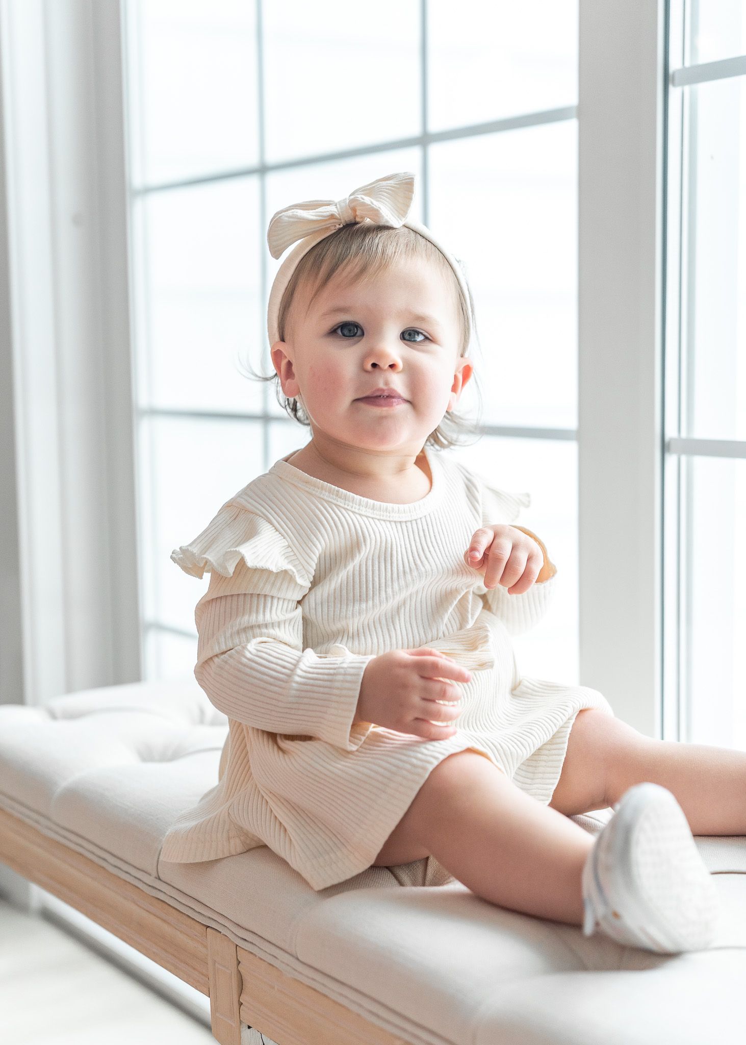 A baby in a cream-colored knit sweater sits on a wooden bench near a bright window.