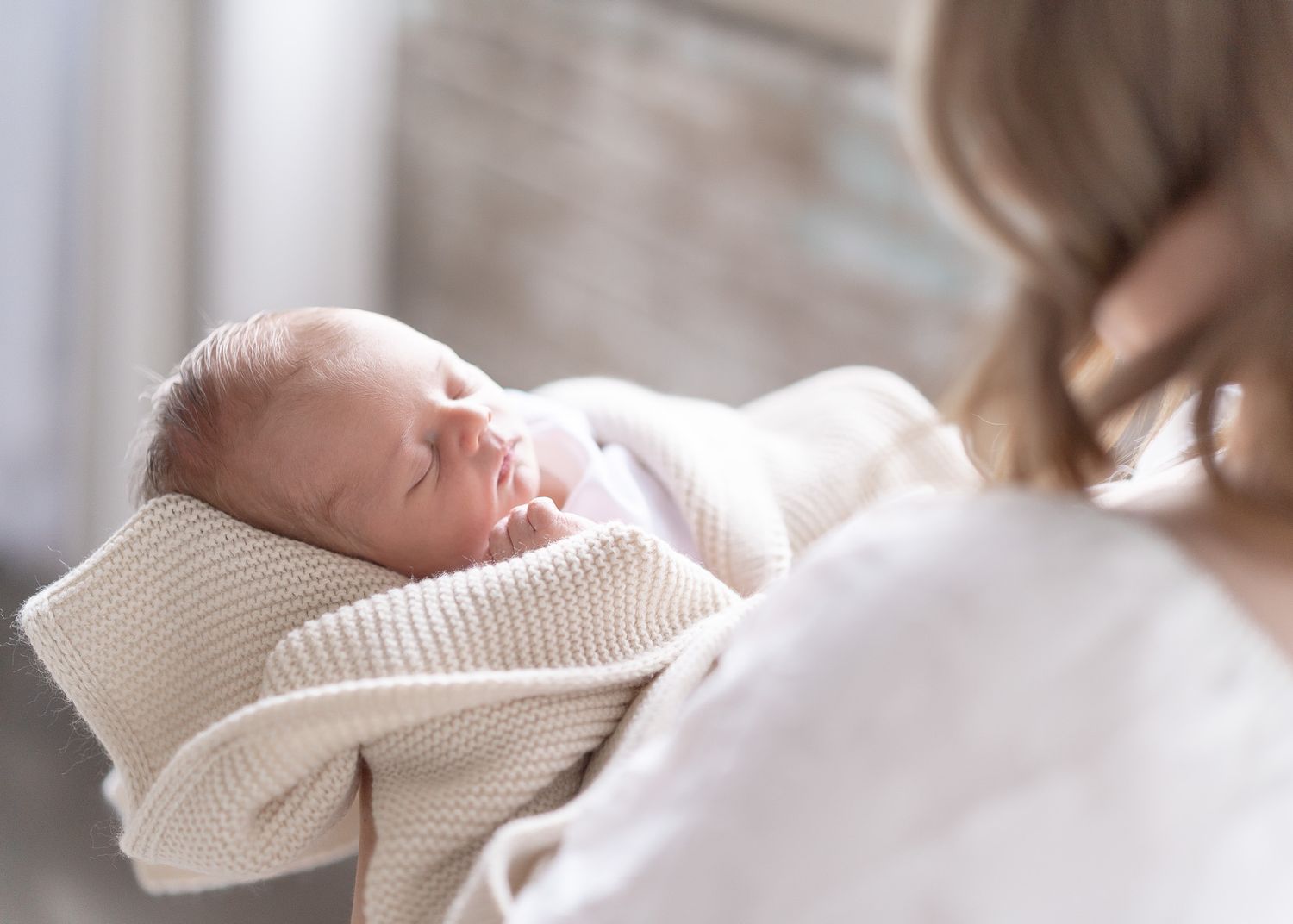 A sleeping newborn baby wrapped in a white knit blanket rests peacefully on a soft surface.