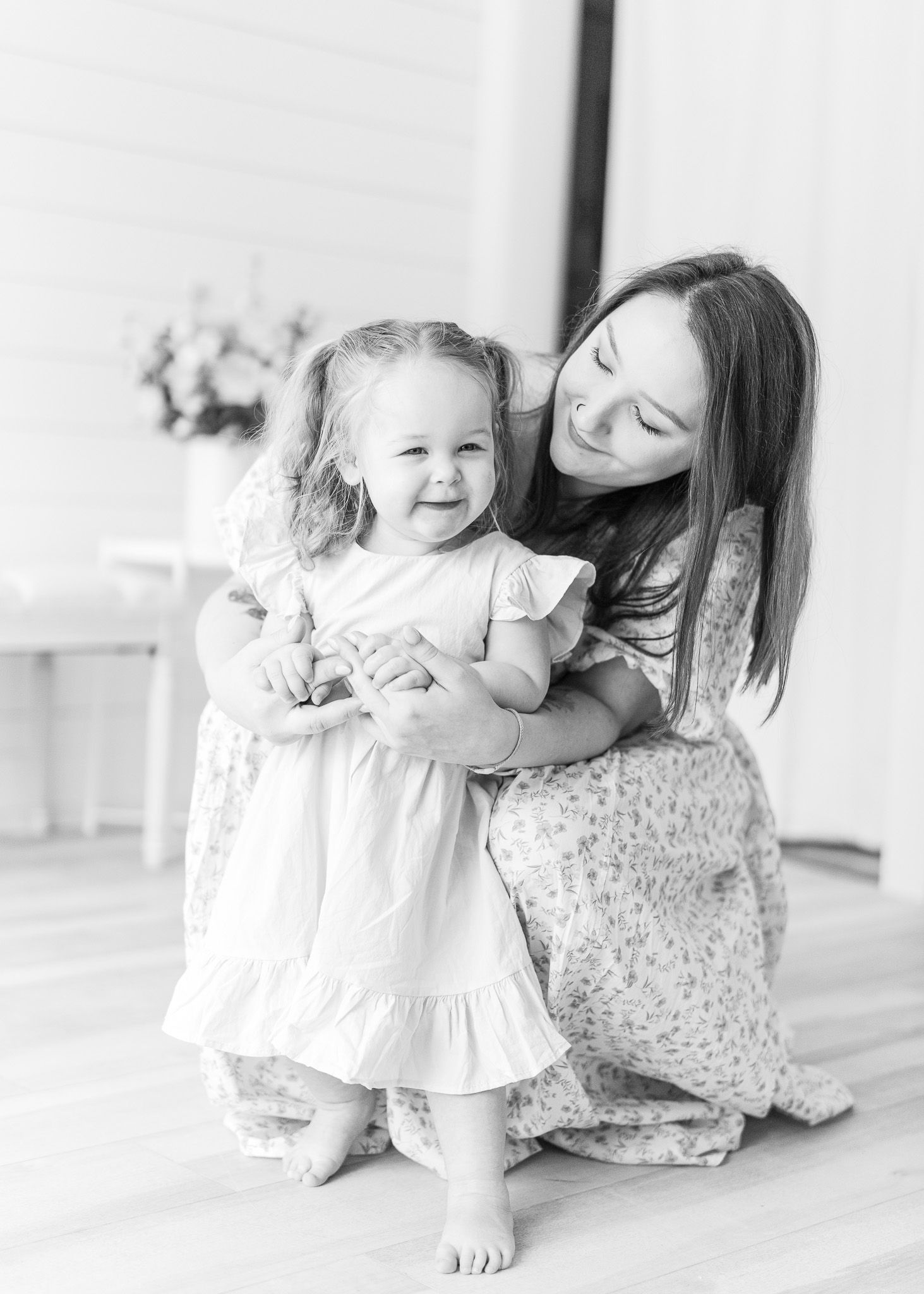 A sweet black and white portrait of a parent and child embracing on a hardwood floor in matching floral dresses.