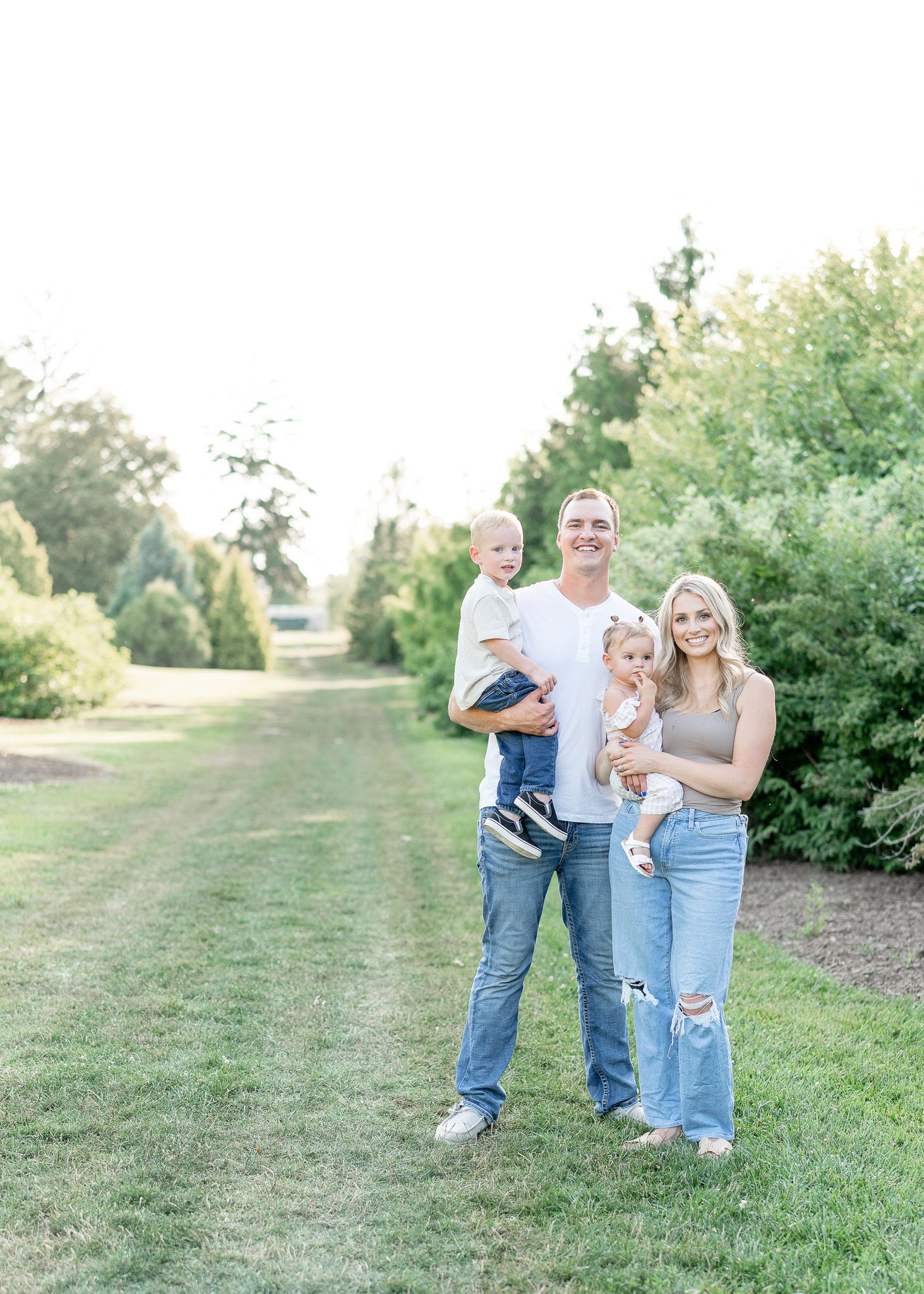 A family stands together on a grassy path lined with green bushes on a bright summer day.