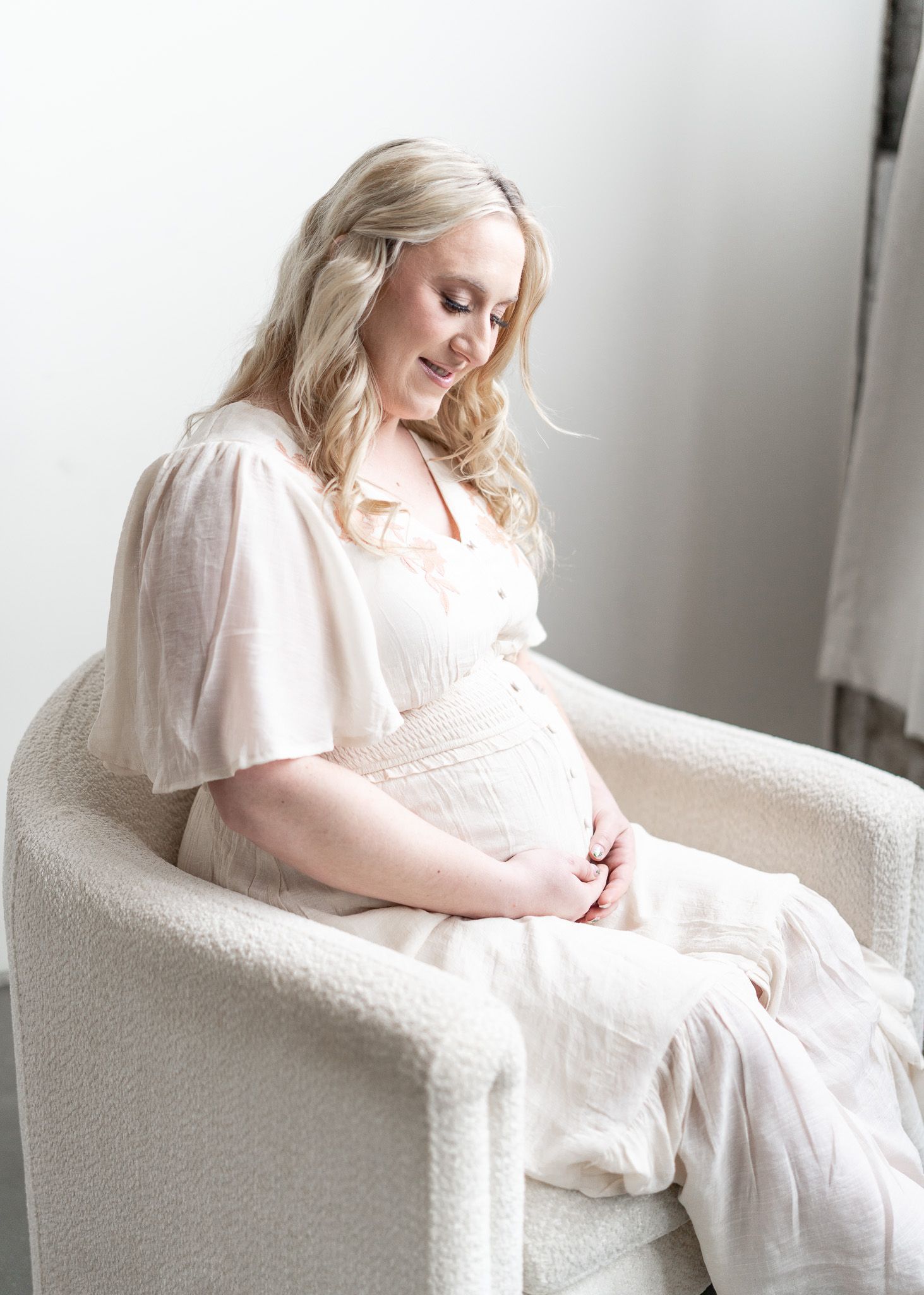 A woman in a soft white dress sits peacefully on a cream-colored armchair in a bright, minimalist setting.