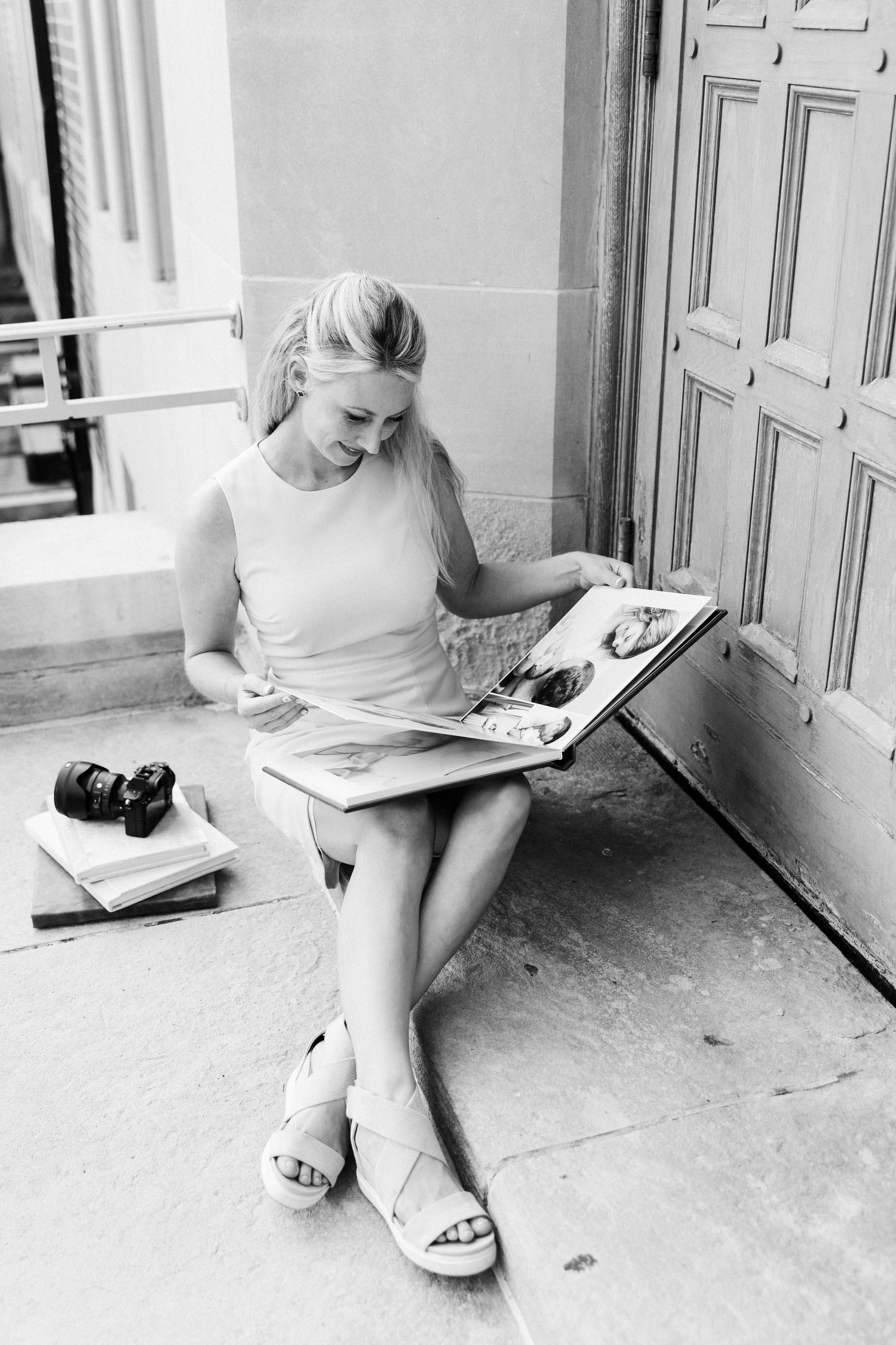Black and white photo of a blonde woman sitting on steps, reading a magazine with a camera beside her.