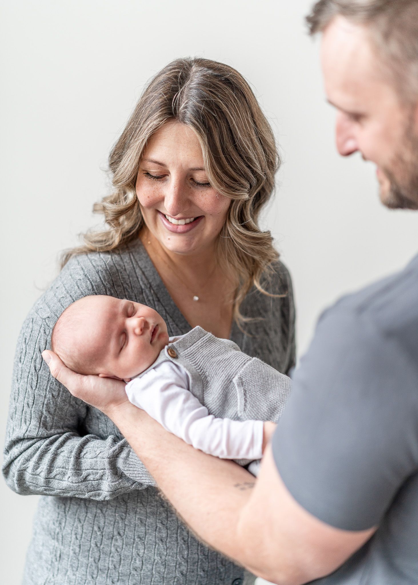 Parents in grey sweaters lovingly admire their newborn baby in a white onesie during an intimate family moment.