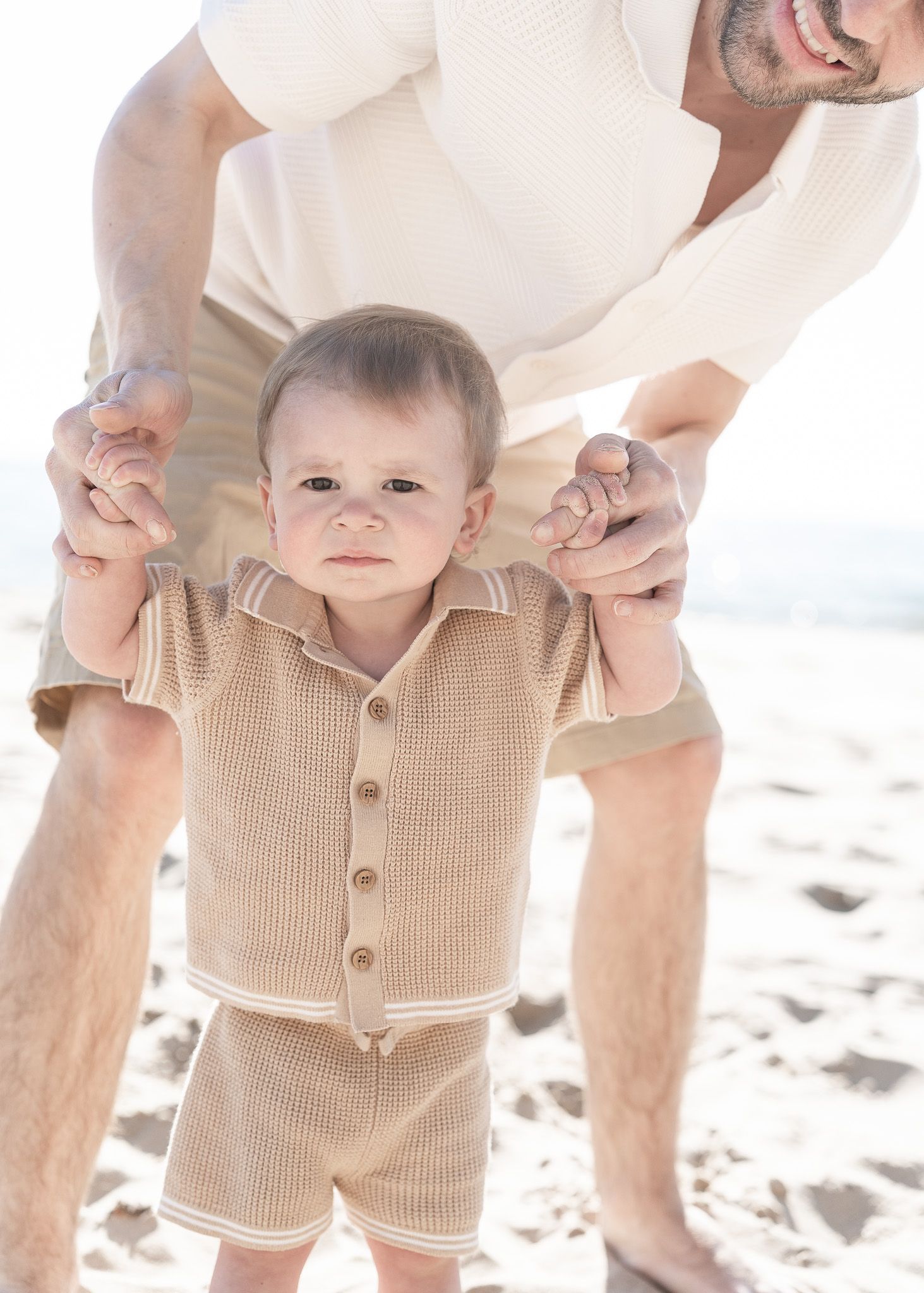 Adorable baby in a beige knit romper crawls on sandy beach while adults hold their hands for support.