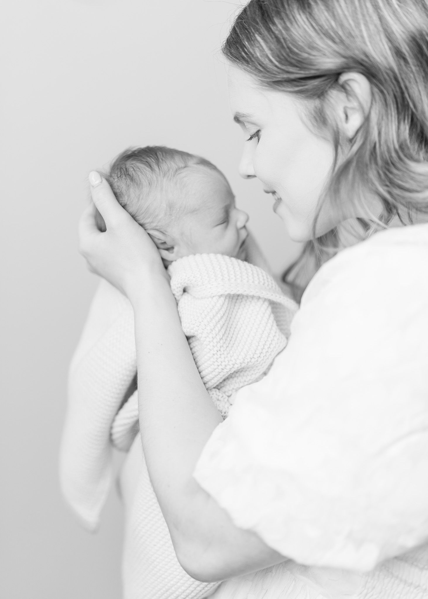 A mother in white gently holds and kisses a sleeping newborn baby in a tender black and white photograph.