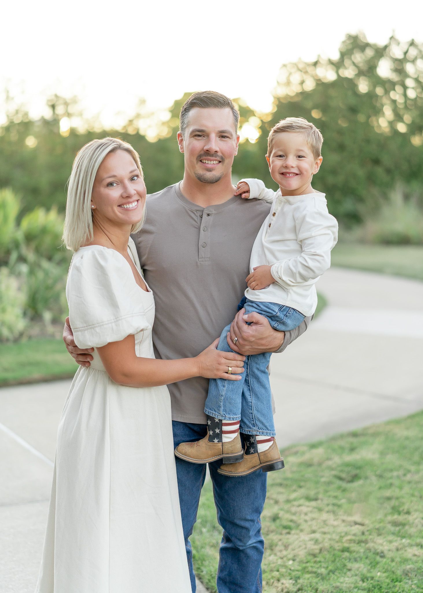A family poses together outdoors on a paved path during golden hour, wearing coordinated casual outfits in white and gray.