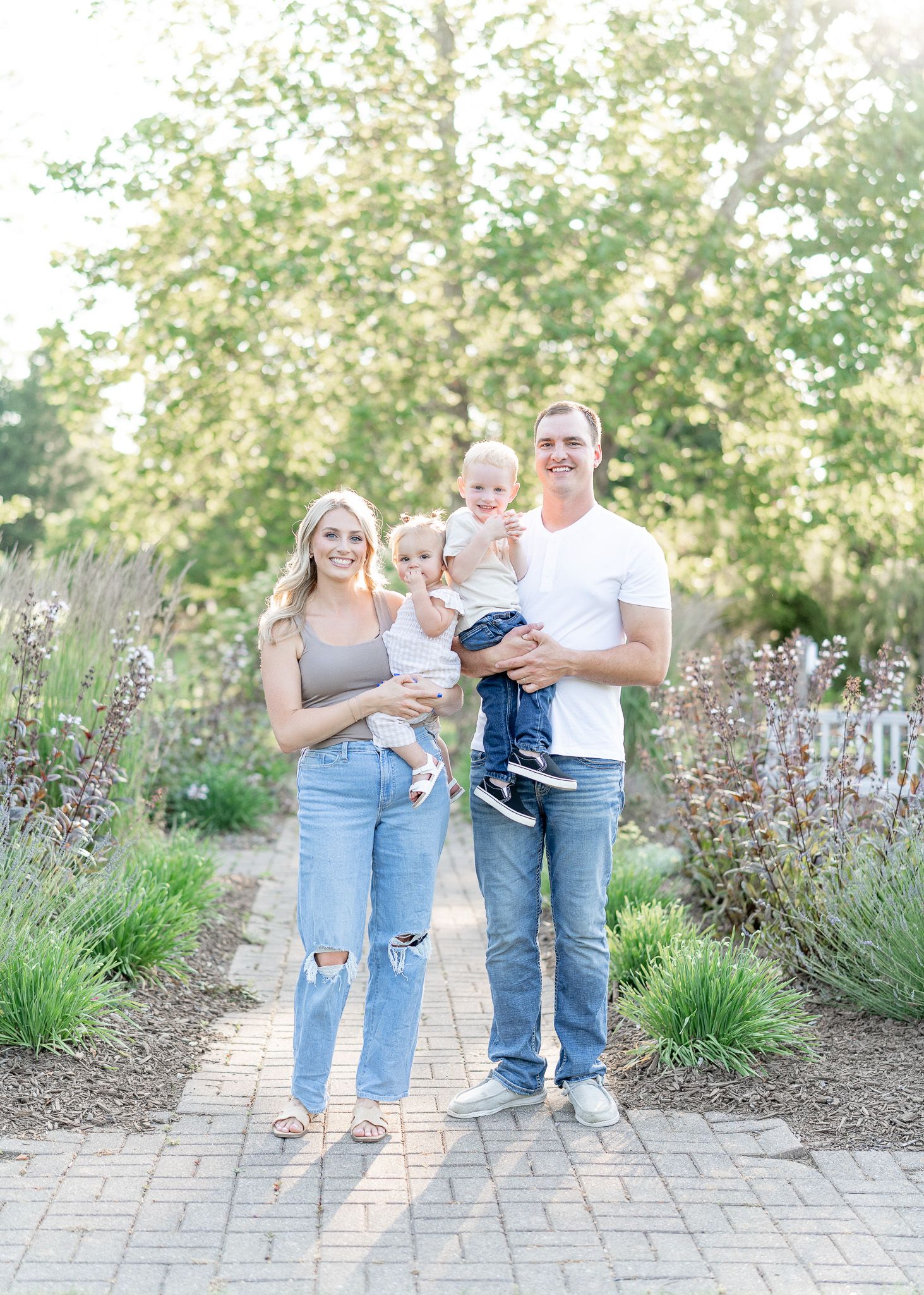 A family stands together on a paved garden path with blooming purple flowers and greenery in the background.