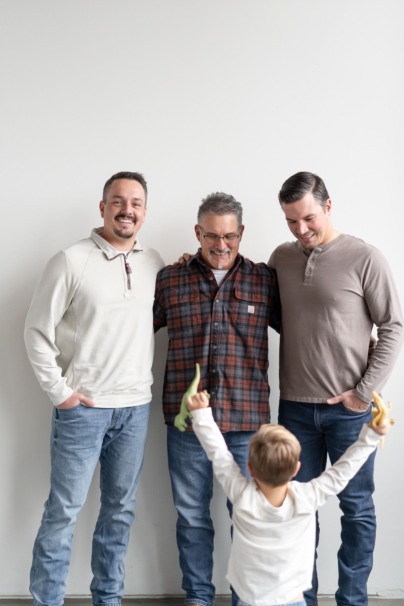 Three adults in casual attire stand together against a white wall while a small child in white raises their arms playfully.