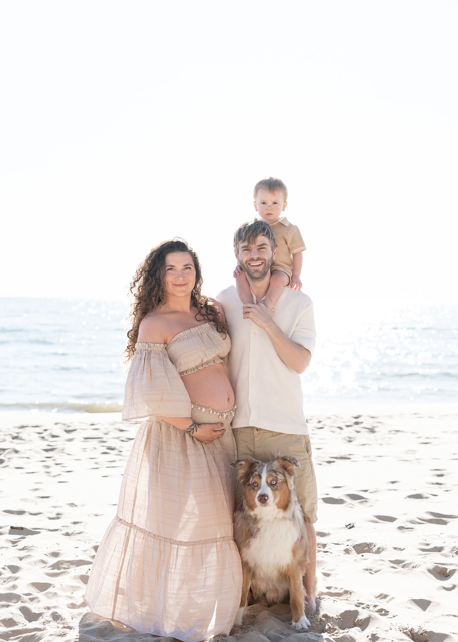 A family and their Australian Shepherd dog pose together for photos on a sunny beach in neutral-colored outfits.