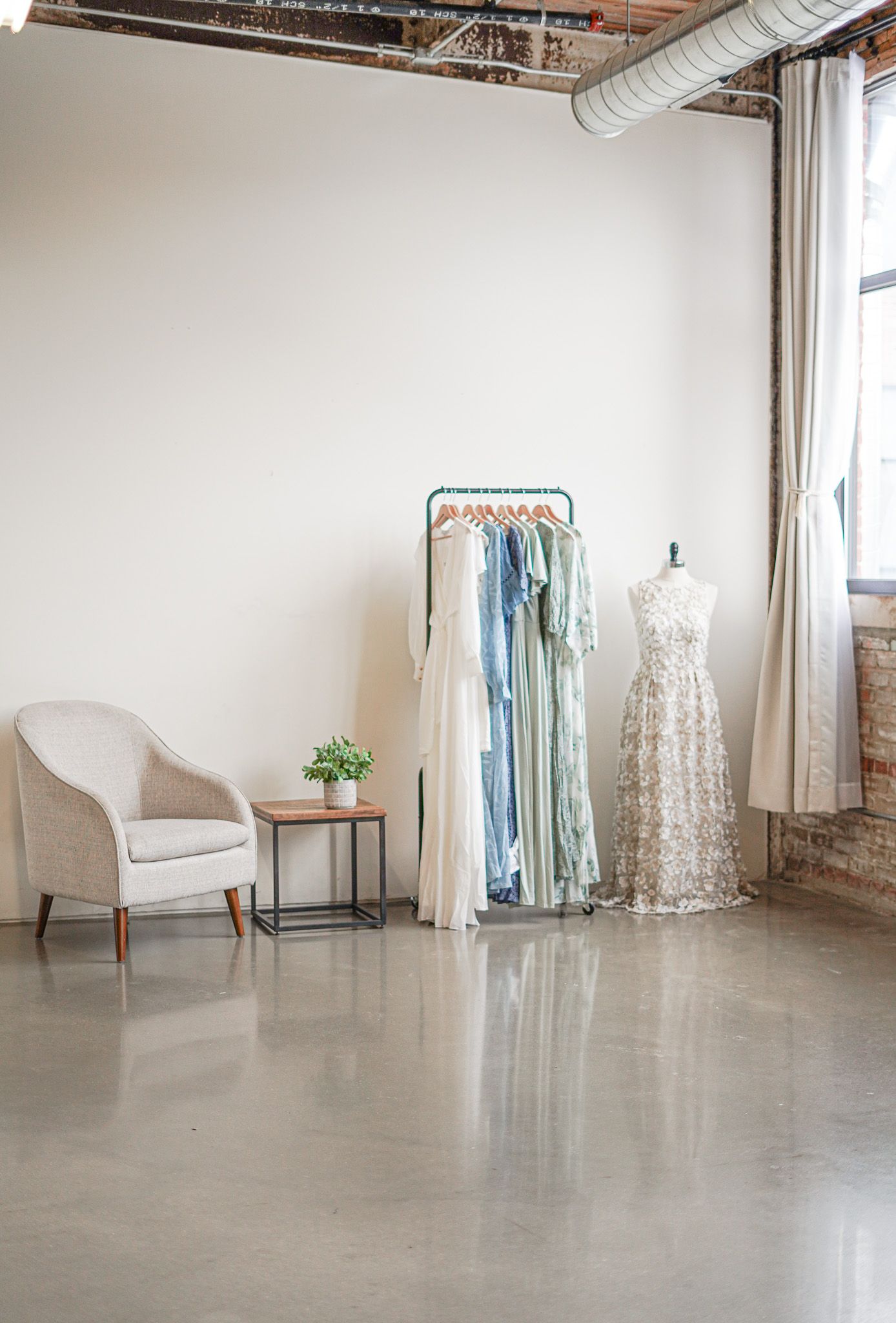 A minimalist bridal boutique interior with a white armchair, clothing rack of pastel dresses, and a mannequin in lace.
