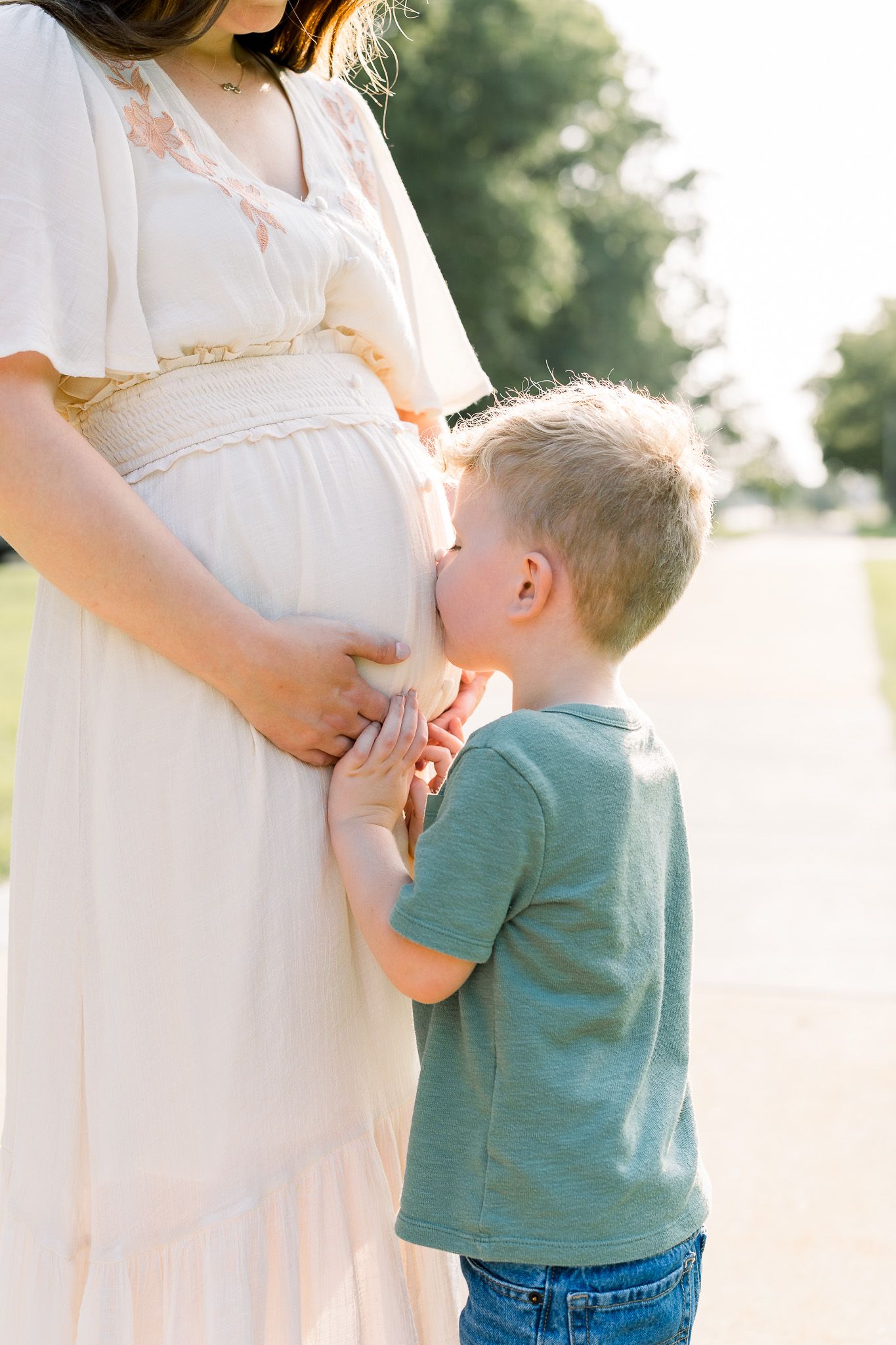 A young boy kisses his pregnant mother's belly outdoors in warm sunlight, wearing a teal shirt and jeans.