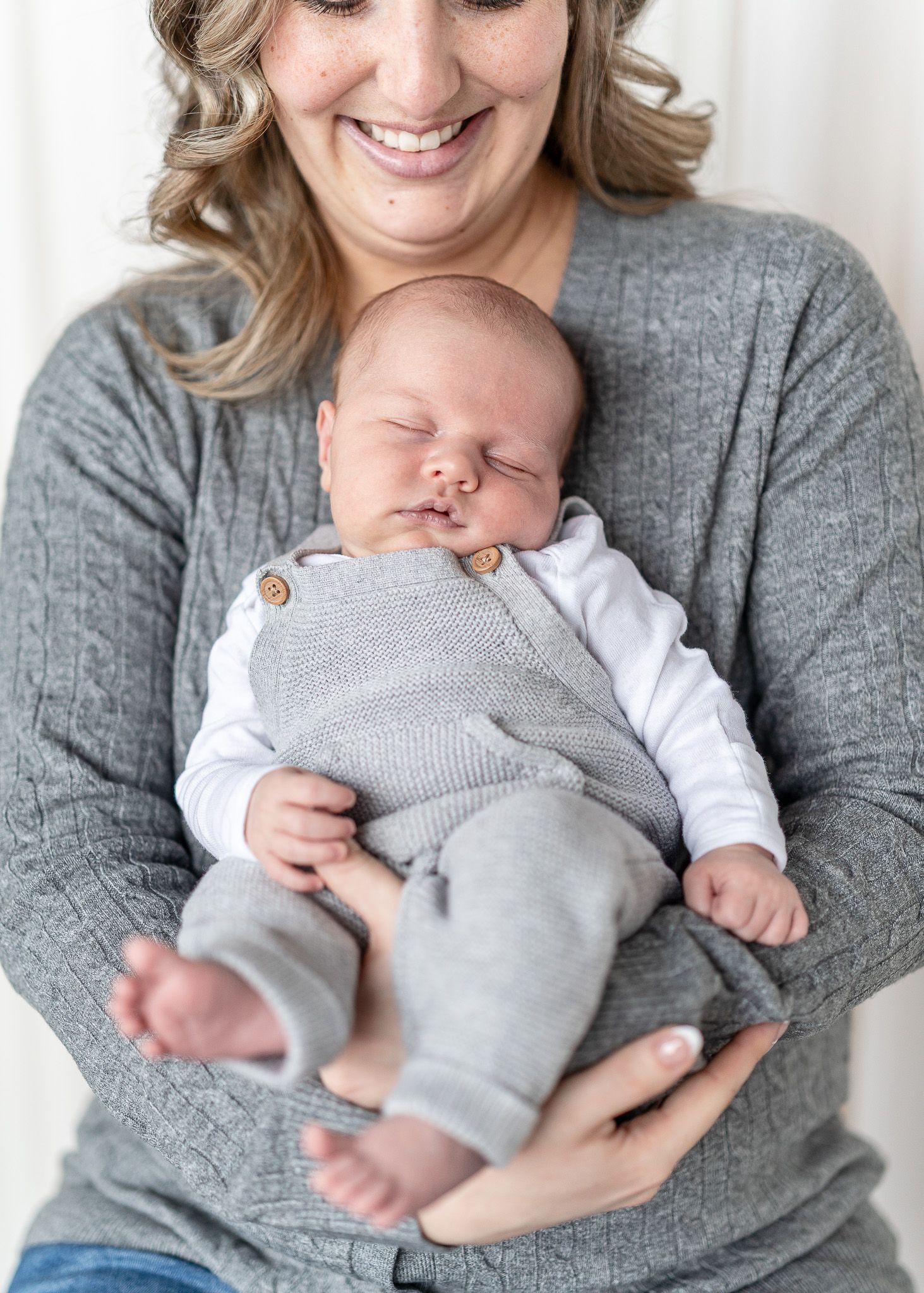 A sleeping newborn baby wearing gray pants and white shirt is cradled in someone's arms wearing a gray sweater.