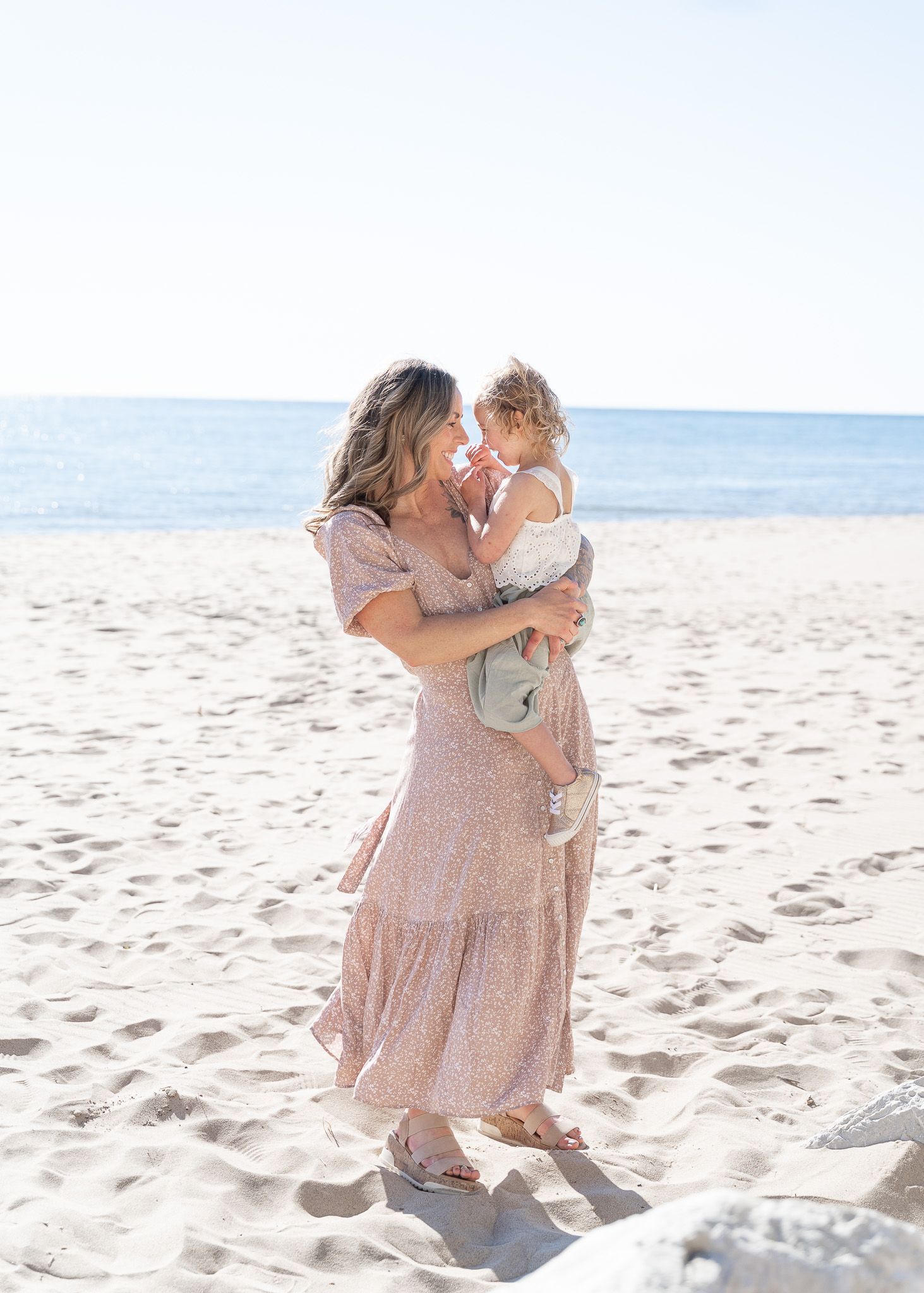 Mother and child in matching pink dresses share tender moments while playing on a sunny beach.