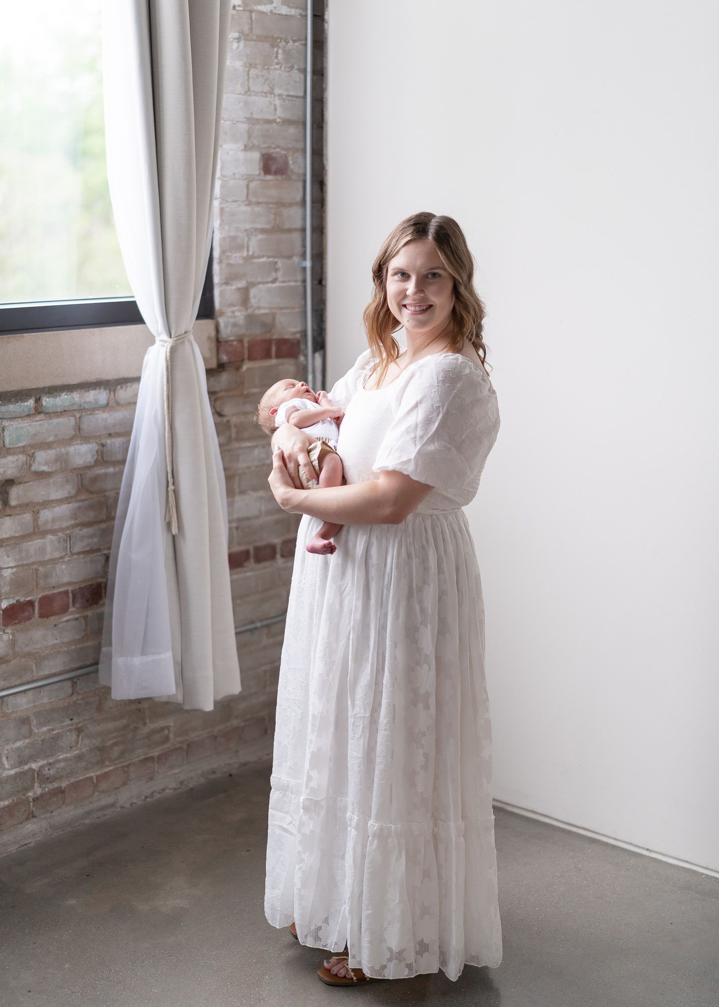 A figure in a flowing white lace dress stands next to a window in a room with exposed brick walls.