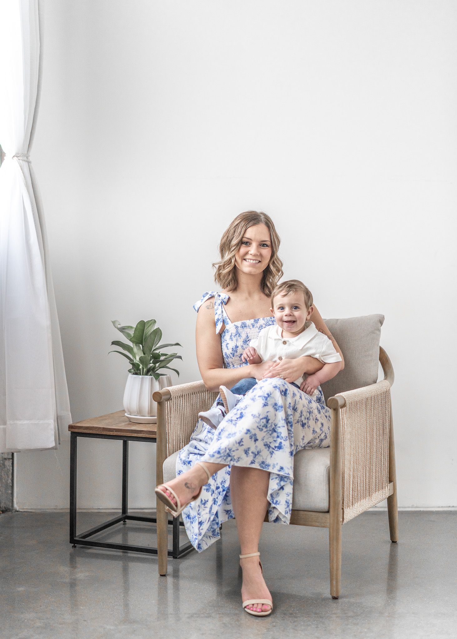 A woman in a blue floral dress sits in a rattan chair with a baby in a bright white studio setting.