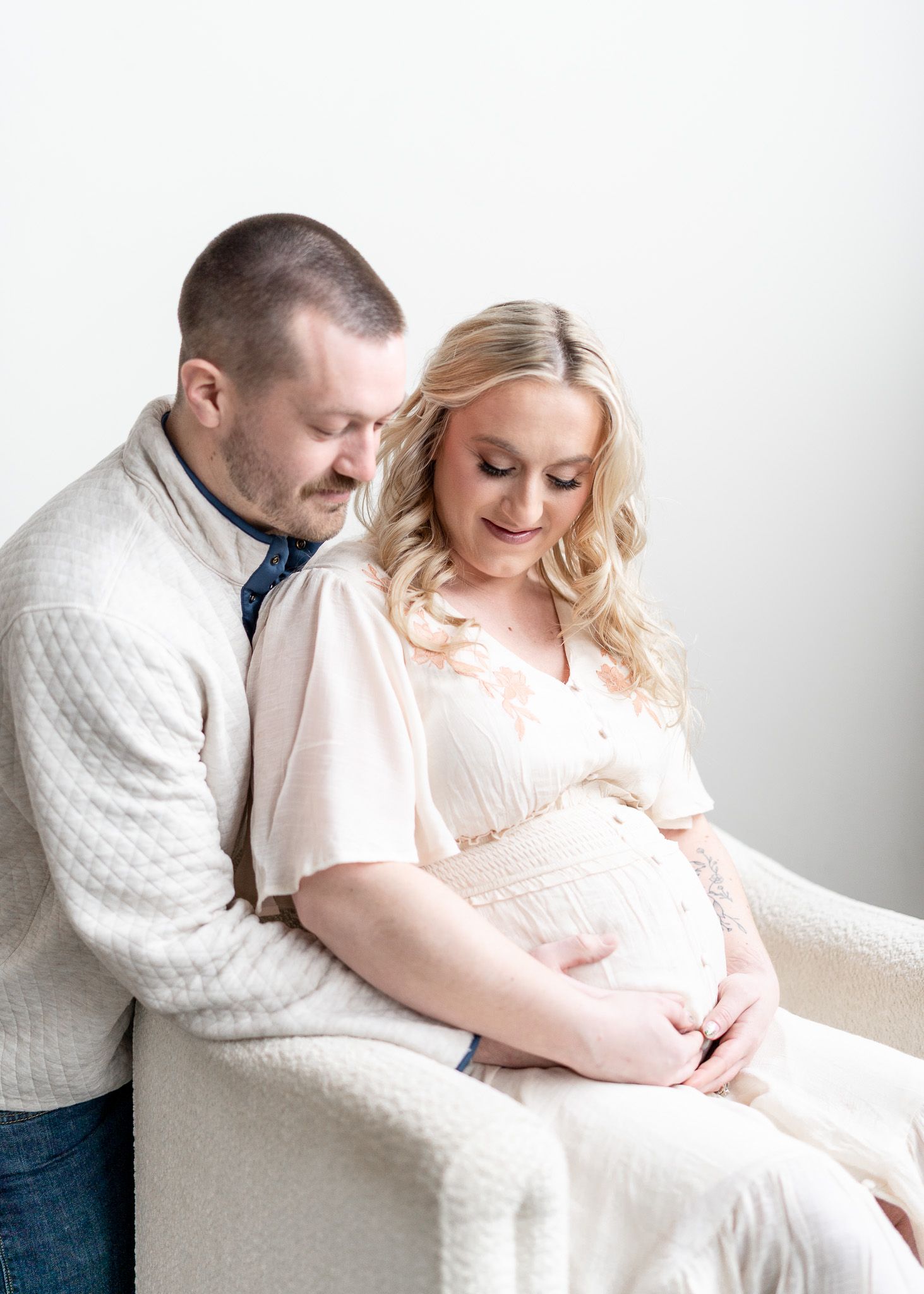 A couple in white clothing share an intimate moment while sitting together during a maternity photo session.