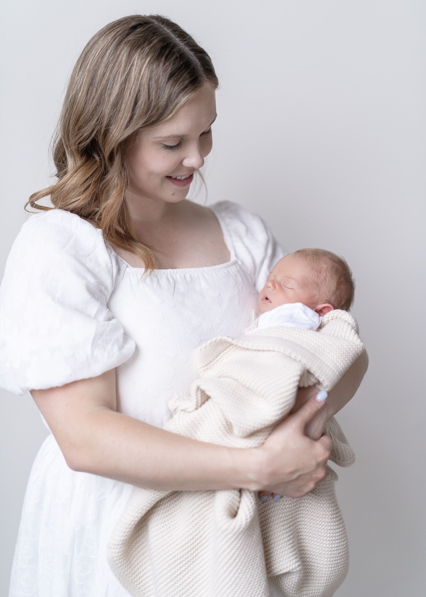 A tender moment captured in soft lighting as a mother in a white dress cradles her sleeping newborn wrapped in cream blankets.