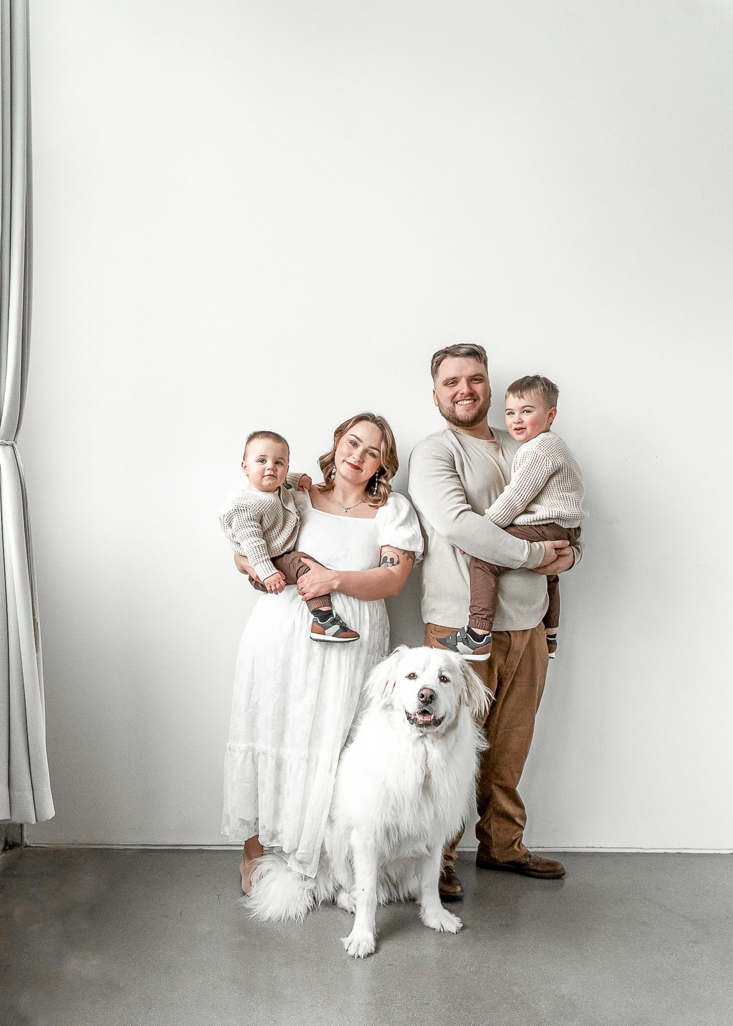 Family in white outfits poses playfully with their large white dog in a minimalist studio setting.