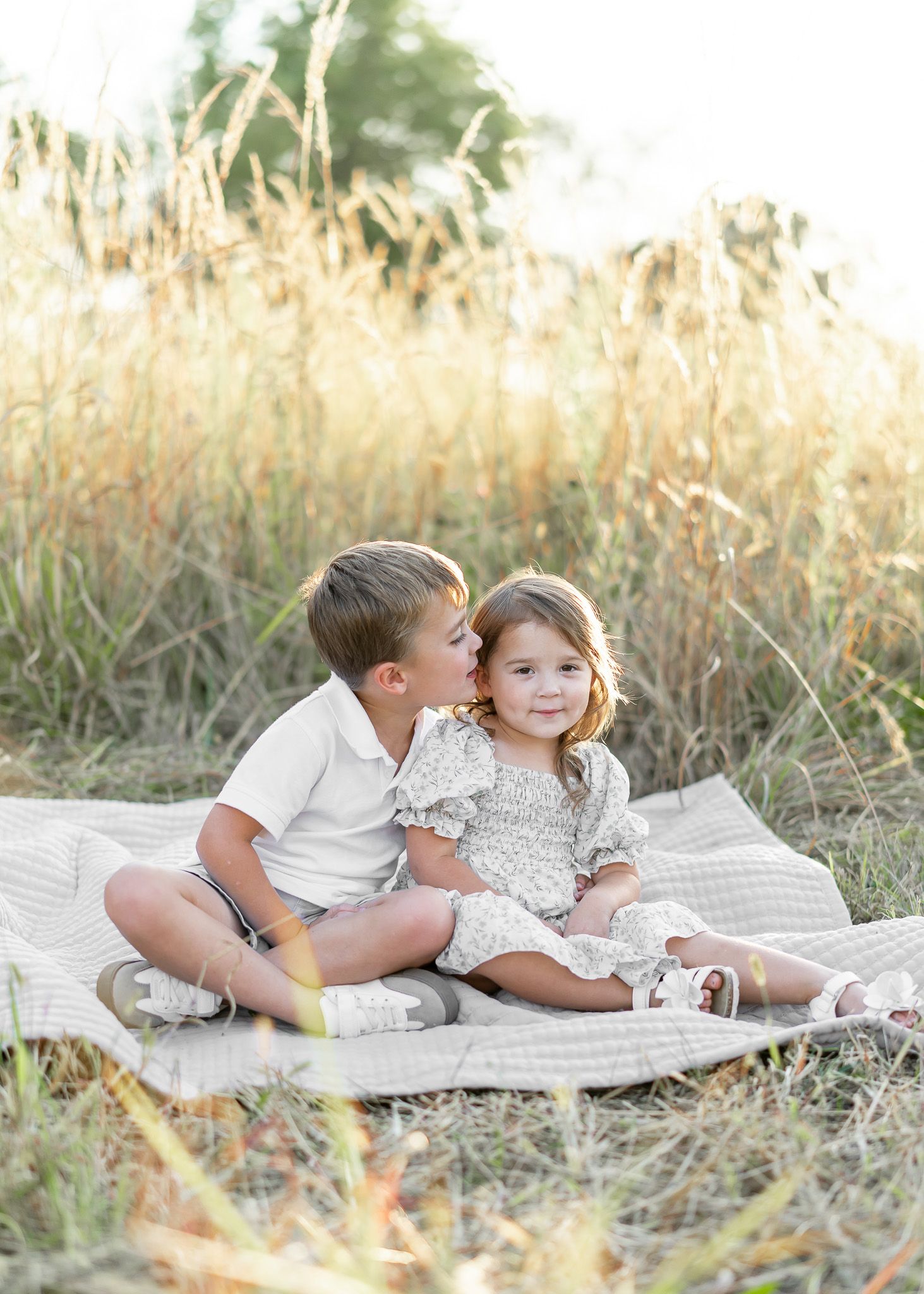Two young children sit together on a white blanket in a field of tall golden grass during sunset.